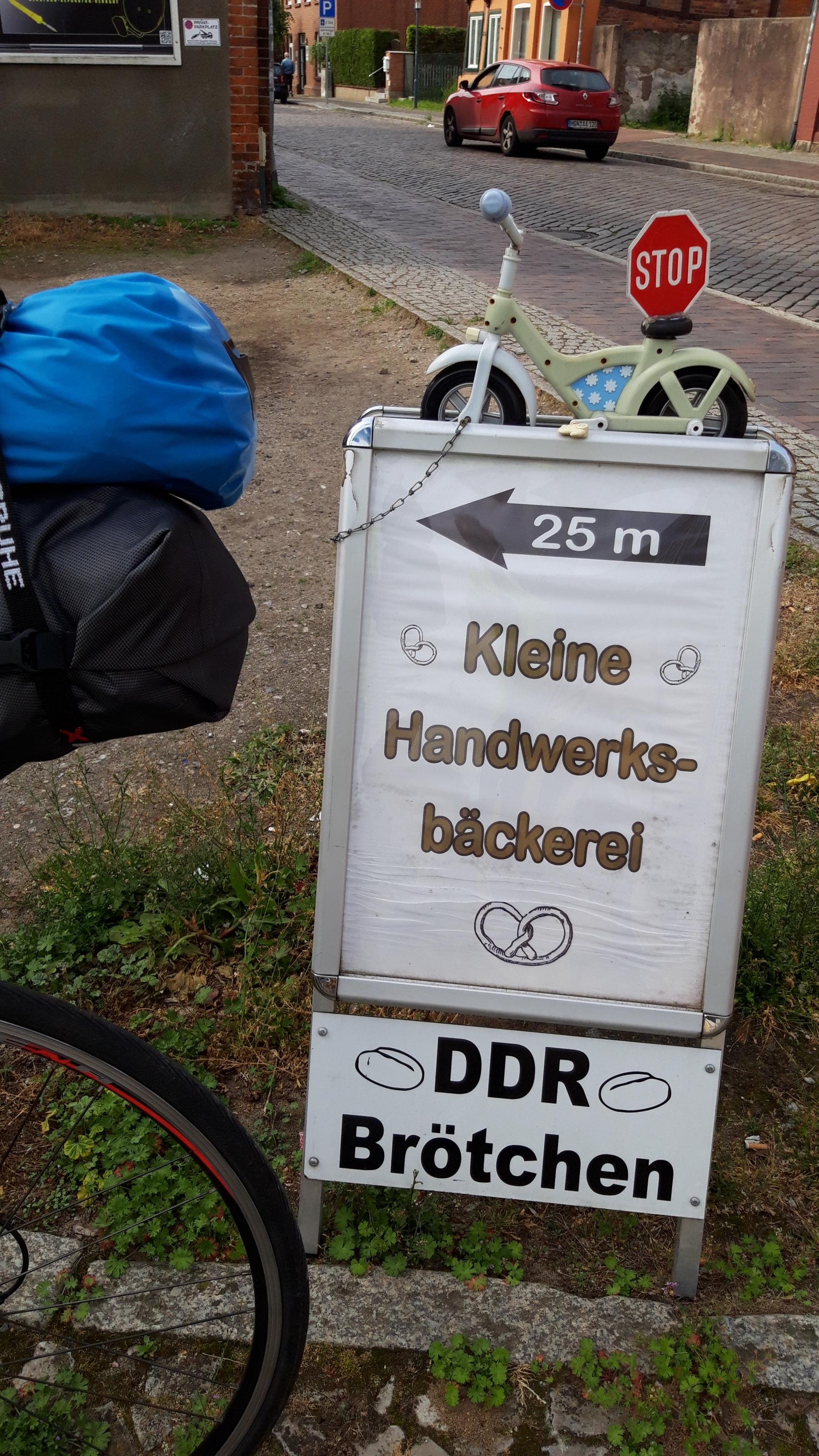 Das Vehikel des RadelndenReporters parkt neben einem Hinweisschild, auf dem zu lesen ist „DDR-Brötchen, Kleine Handwerksbäckerei, 25 Meter“.
