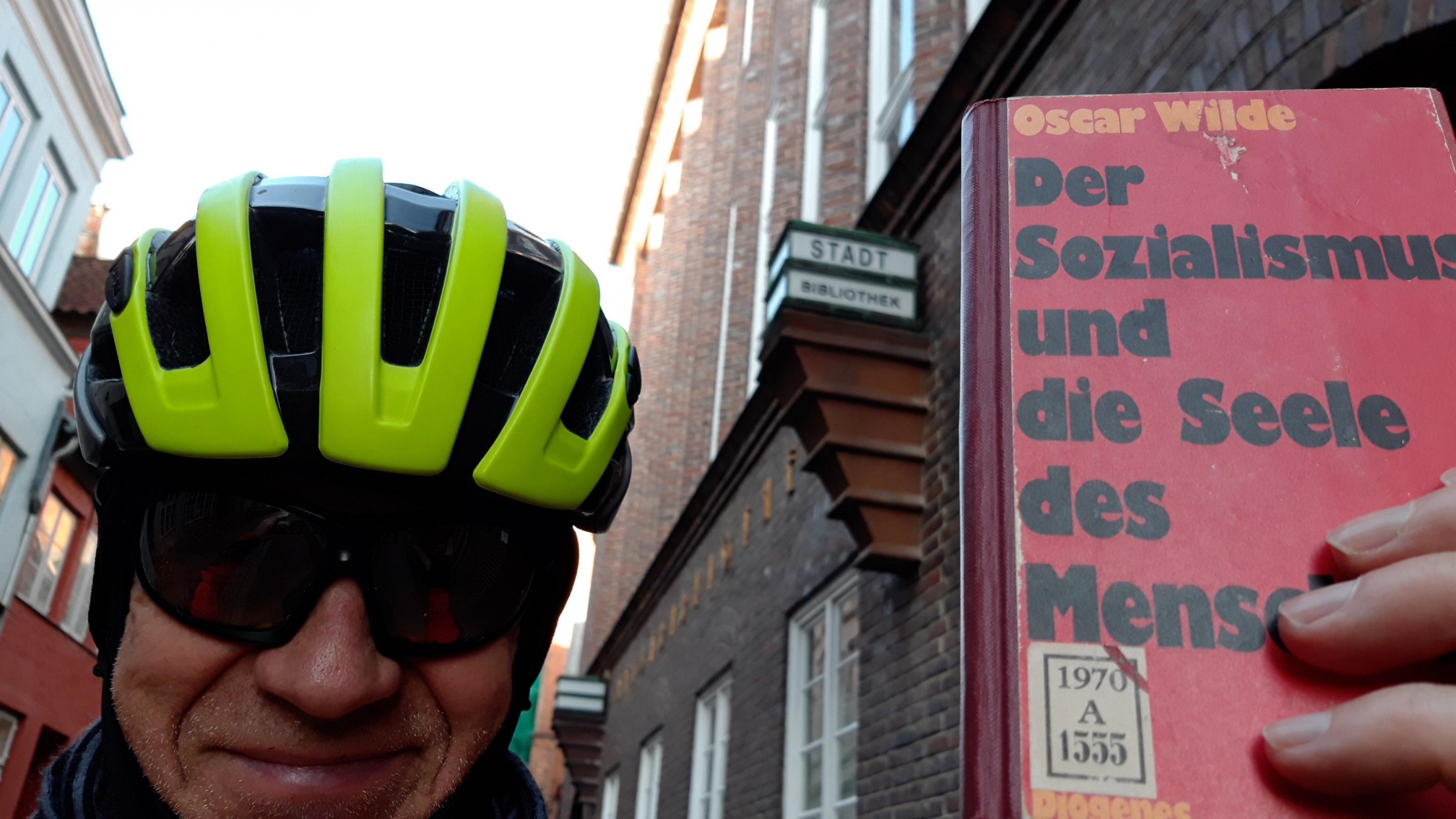 Martin C Roos steht mit Radhelm vor der Stadtbibliothek Lübeck und hält triumphierend das eben ausgeliehene Büchlein in der Linken.