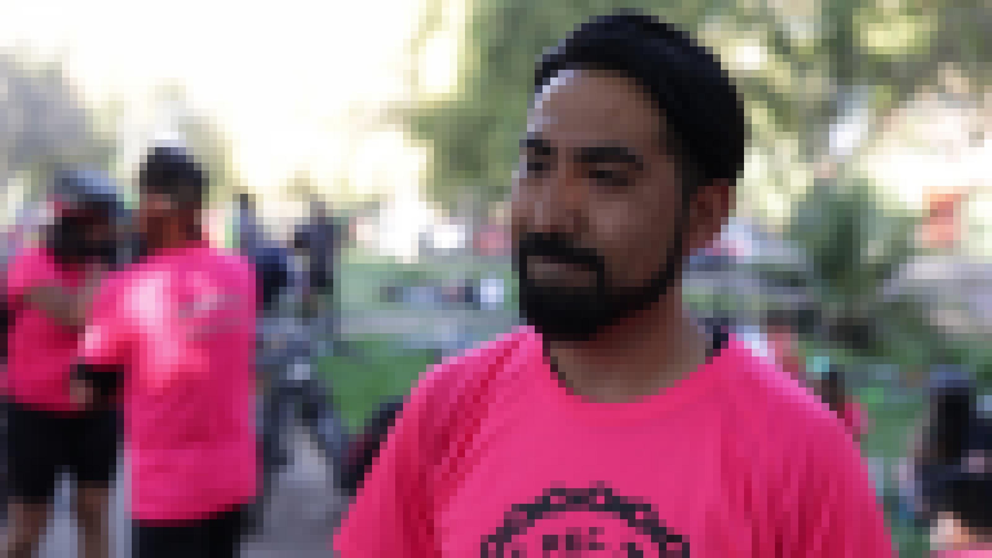 Ein Mann Mitte 30 mit Vollbart und pinken T-Shirt. Im Hintergrund sind zwei weitere Menschen mit dem T-Shirt und Radlerhosen zu erkennen.