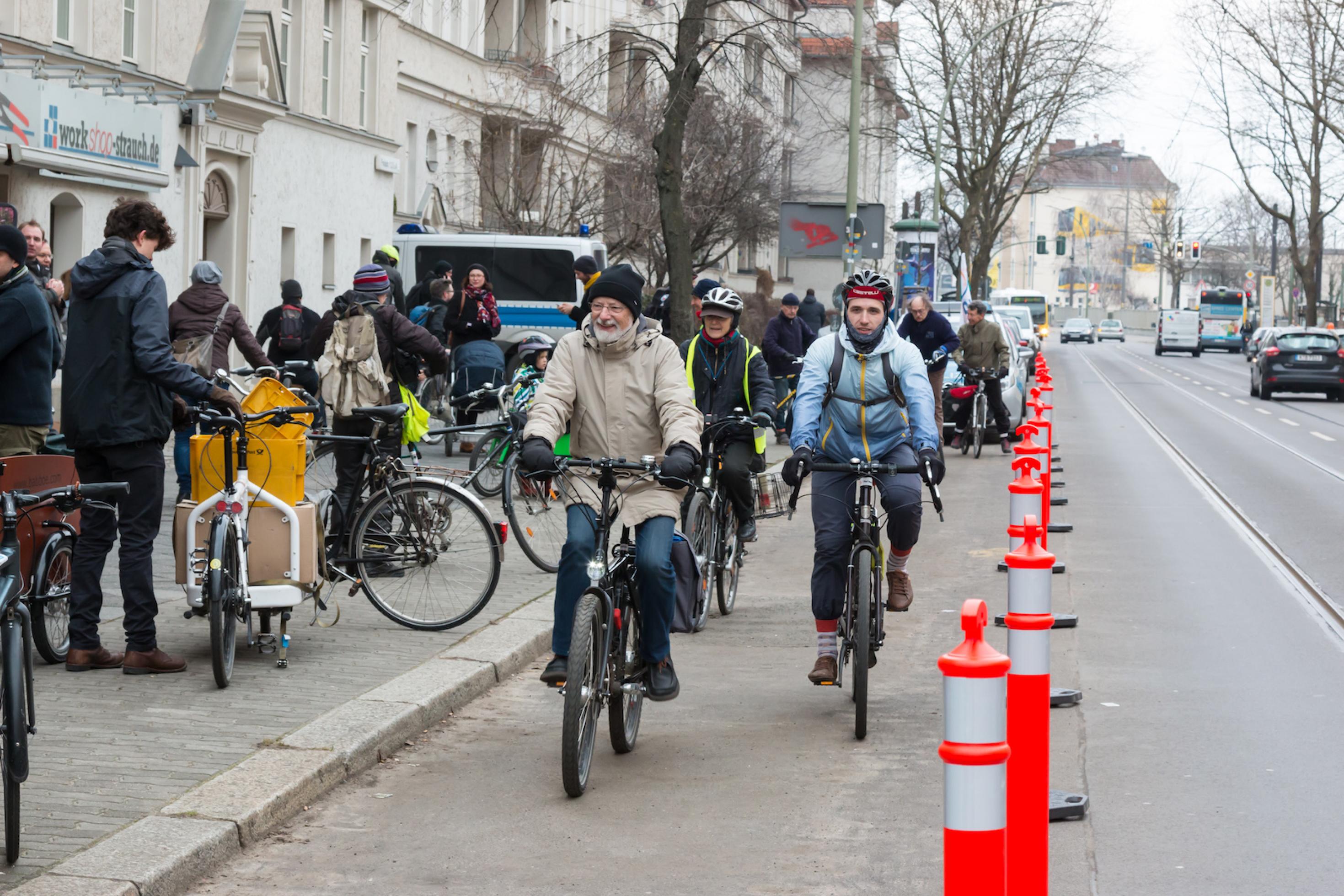 Mehrere Radfahrer fahren in Richtung Kamera auf einer gesicherten Spur. Auf dem Bürgersteig stehen weitere Menschen mit ihren Fahrrädern.