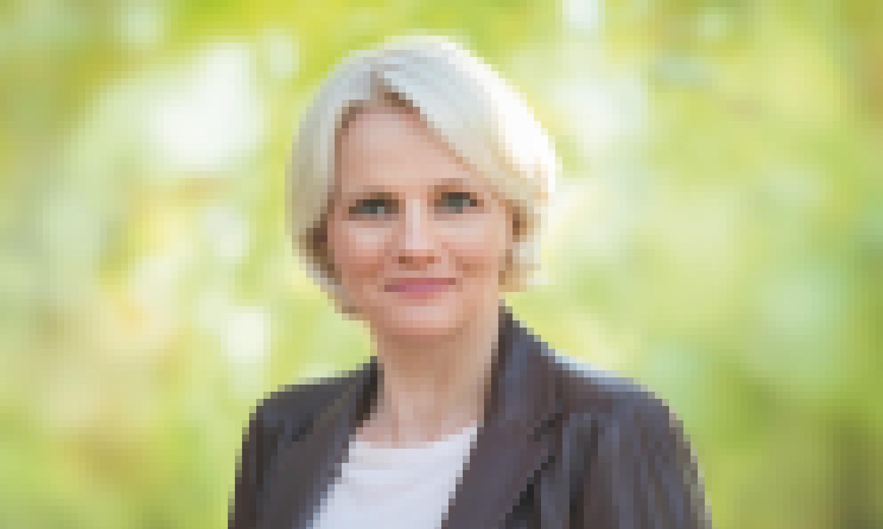 Regine Günther hat hellblonde Haare, trägt Perlenohrringe, ein helles Shirt und eine dunkelbraune Lederjacke. Sie steht vor einem hellgrünen verschwommenen Hintergrund