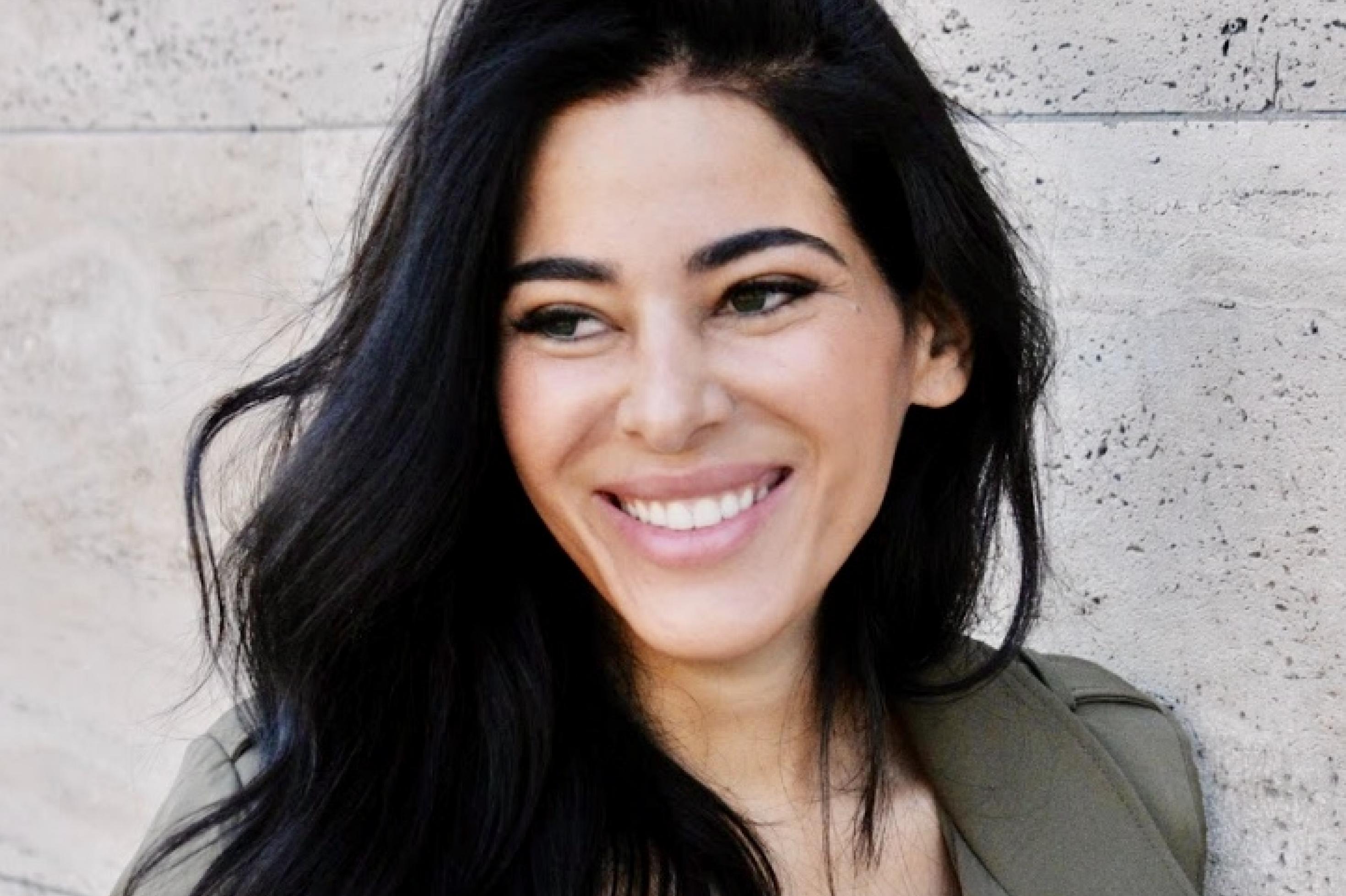 Porträtfoto von Samira El Ouassil: Frau mit dunklem langen Haar schaut lächelnd in die Kamera, im Hintergrund eine beige Steinmauer