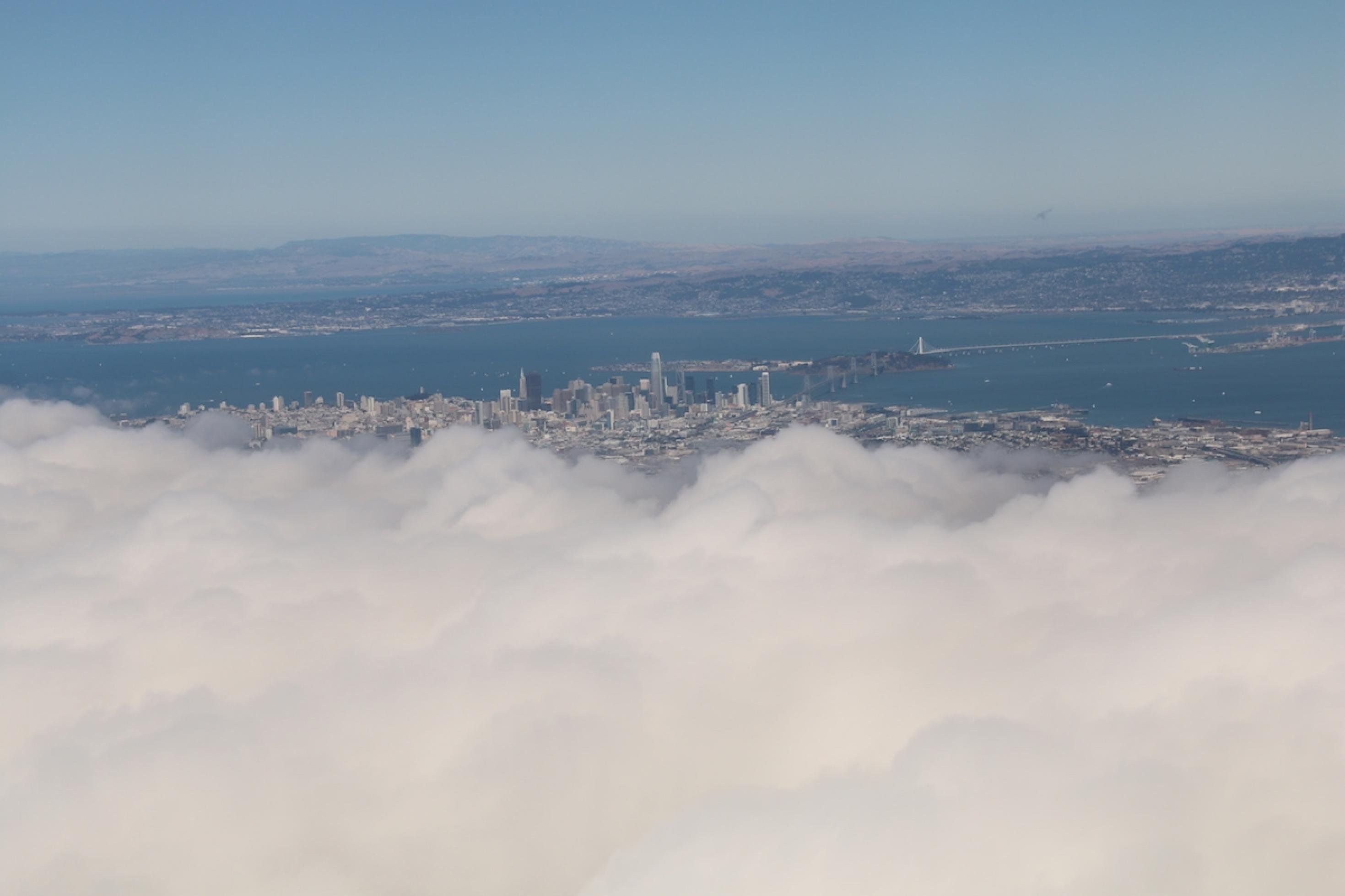 Blick aus dem Flugzeug-Fenster. Durch eine Wolkenlücke erkennt man die Innenstadt von San Francisco Oakland-Bay-Brücke. 
Abschied von San Francisco mit Blick auf die Skyline und die Oakland-Bay-Bridge. Bleiben die Versprechen, die in der Stadt abgegeben wurden, nur wolkige Worte?