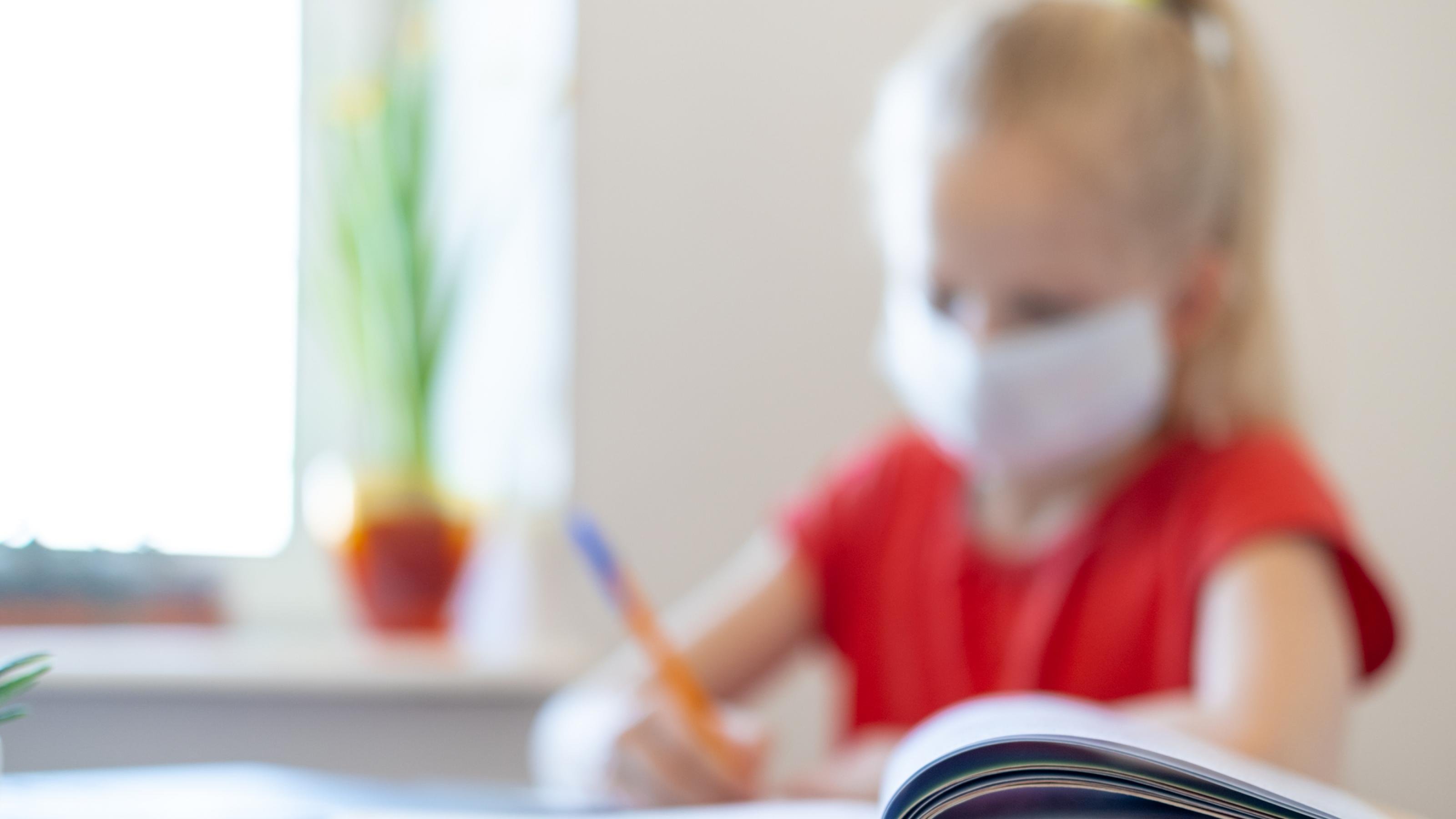 Ein kleines Mädchen sitzt mit Maske an einem Schreibtisch und macht Aufgaben in einem Schulheft.