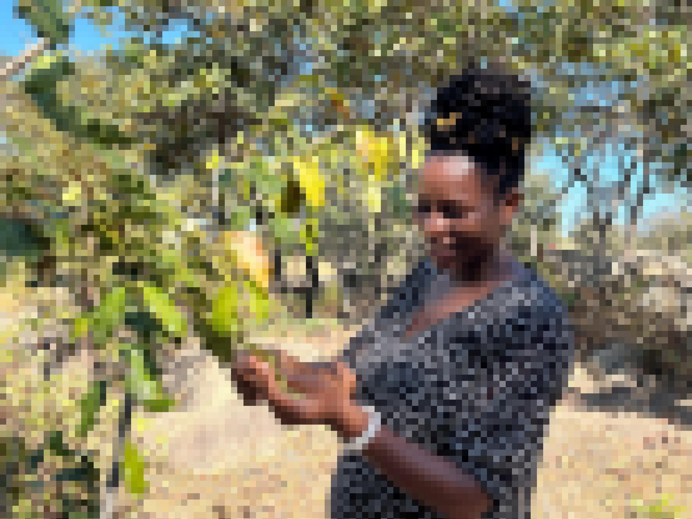 Eine eher junge Frau steht vor einem kleinen Baum, blickt auf ein Blatt (oder Samen?) in ihrer Hand und lächelt breit.