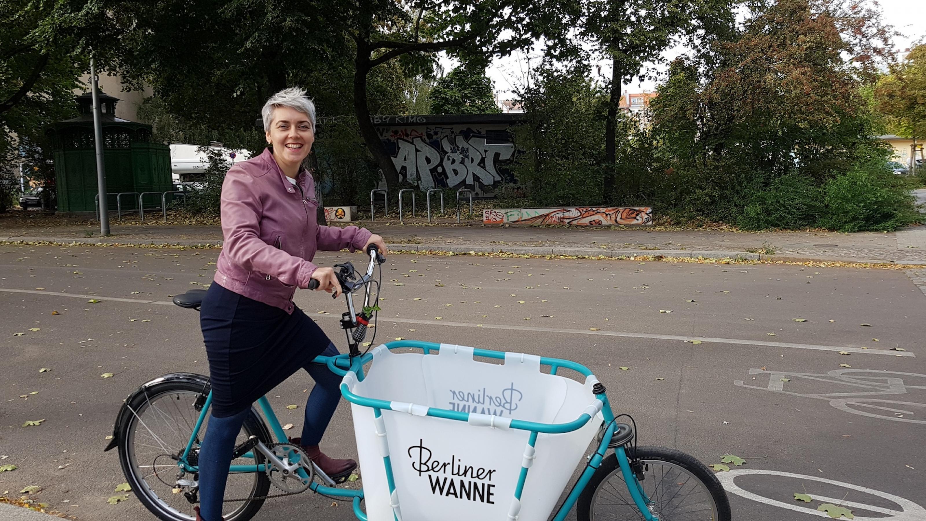 Die Mobilitätsforscherin Sophia Becker steht mit einem Lastenrad auf einer zweispurigen Straße. Das Lastenrad hat einen türkisfahrbenenn Rahmen und vor dem Lenker eine weiße Box. In türkisfarbener Schrift  steht „Berliner Wanne“ auf der Lastenradbox.