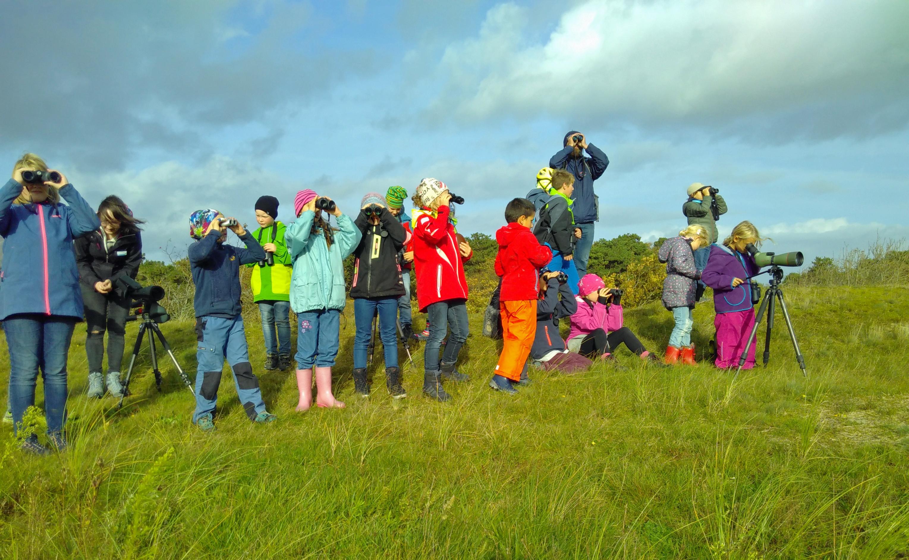 Eine Gruppe von rund 15 Kindern in bunten Anoraks steht auf einer Wiese und blickt durch Ferngläser und ein Spektiv