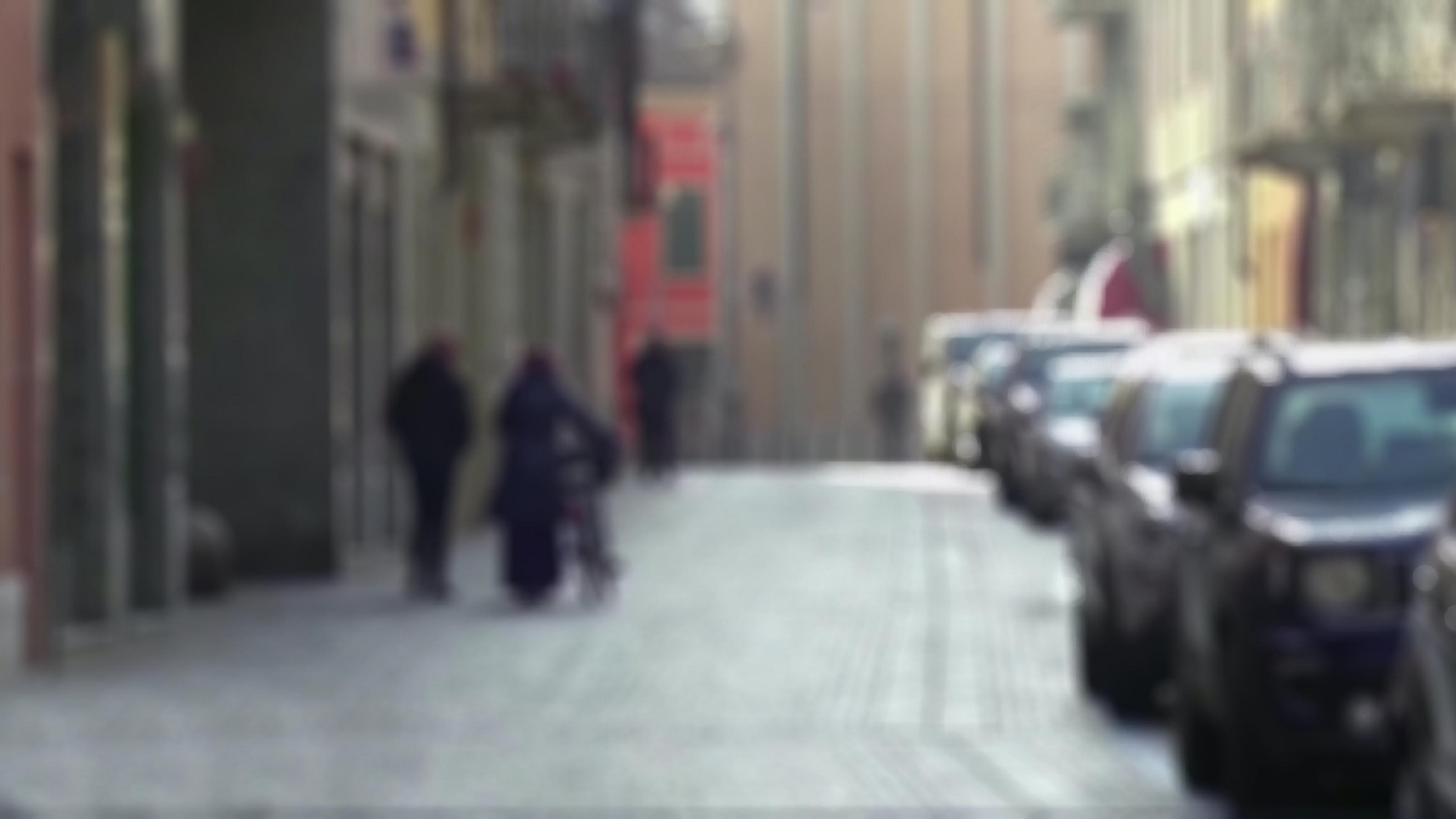 Das Bild ist unscharf und zeigt eine Straße in der links zwei Fußgänger laufen, einer von ihnen schiebt sein Fahrrad, rechts parken Autos