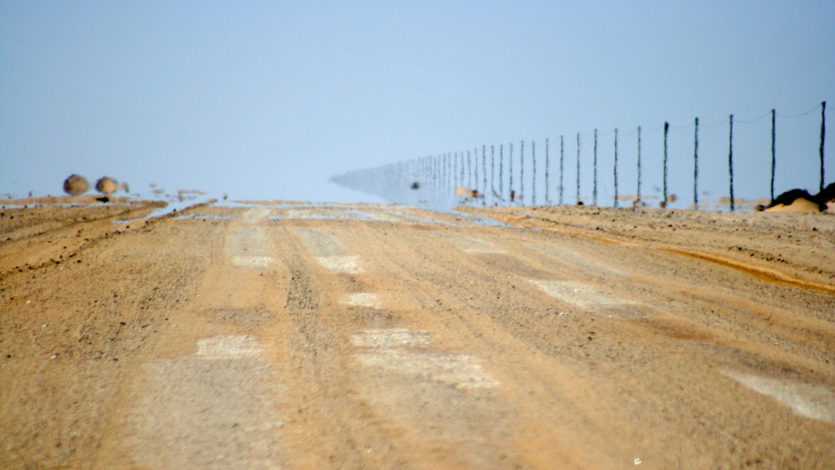 Eine Sandpiste, rechts steht eine Reihe von Strommasten. Weiter hinten verschwimmen Wüstenboden und Masten wegen einer Luftspiegelung mit dem Himmel. Wohin also führt diese Straße?