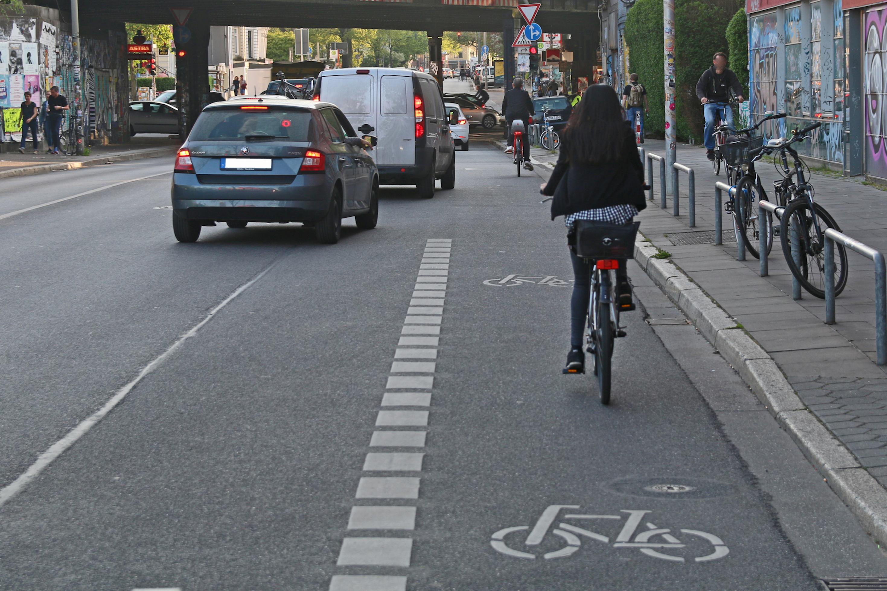 Eine Fahrradfahrerin rollt auf eine rote Ampel zu. Neben ihr zeigt eine gestrichelte Linie an, dass der Fahrradweg endet. Vor ihr ziehen Autos nach rechts, dort fährt ein weiterer Radfahrer. –
… Max-Brauer-Allee an der Sternbrücke. Der Radweg endet, viele Autos wollen rechts abbiegen. Hier wird es oft verdammt eng. …