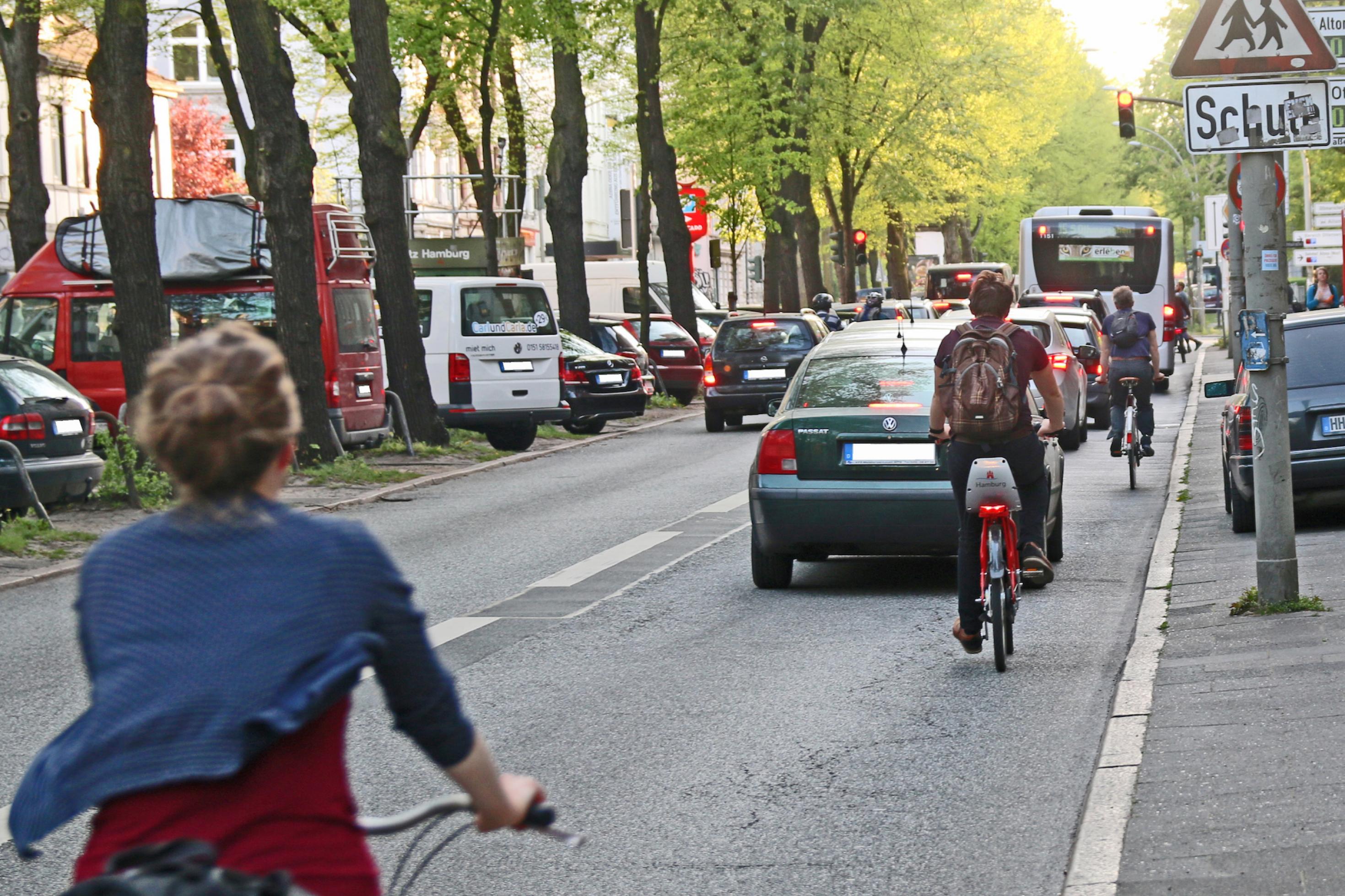 Das Foto zeigt Radfahrer, die sich zwischen einer wartenden Autoschlange und dem Bordstein bewegen. –
… Weiter auf der Max-Brauer-Allee: Fahrräder und Busse und Rechtsabbieger. Die Autoschlange hier ist oft lang, der Abstand zwischen Autos und Bordstein schmal.