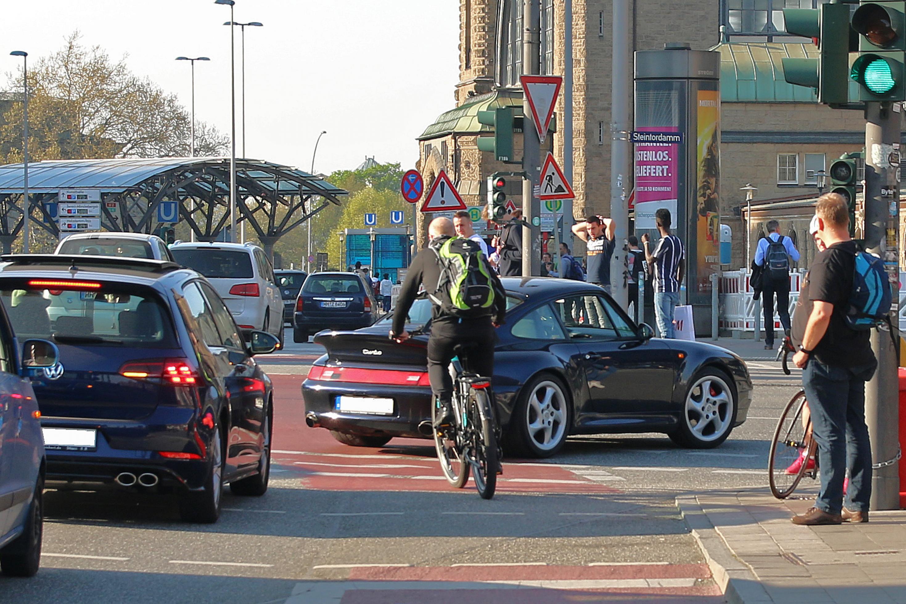 Vor dem Hauptbahnhof: Ein schwarzer Sportwagen steht quer auf dem rot markierten Fahrradweg, ein Mann auf seinem Fahrrad schlängelt sich zwischen Heck des Autos und dem nächsten Wagen in der Schlag vorbei. Straßenszenen in Hamburg: Weil der Porsche beim Rechtsabbiegen den Radweg blockiert, muss der Radfahrer zwischen den Autos Slalom fahren …