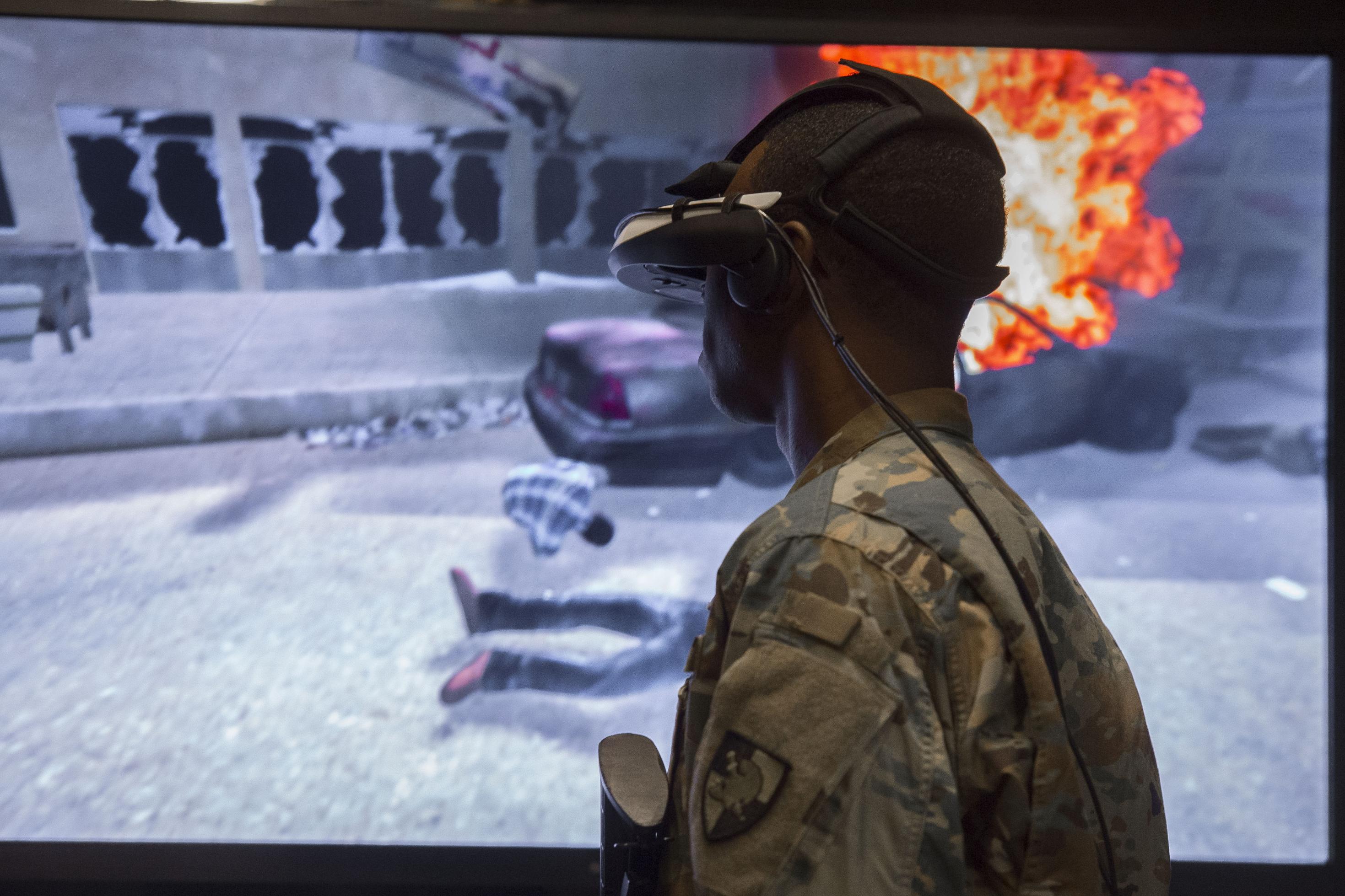 Ein Kadett des US Militärs in Tarn-Kleidung steht mit einem VR-Headset vor einem großen Monitor auf dem die virtuelle Szene eines Bombenanschlages zu sehen ist.