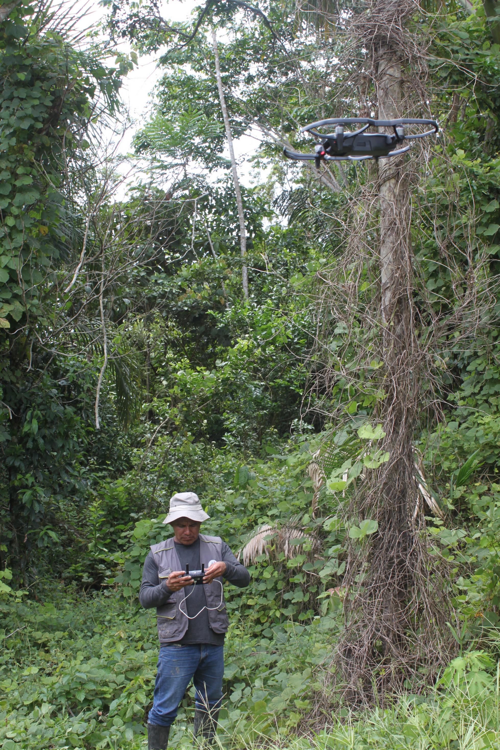 Ein Mann mit Trapperjacke und Hut bedient eine Drohne. Der Mann steht inmitten eines Gebüschs, ist von hohen Bäumen und Gebüsch umgeben.