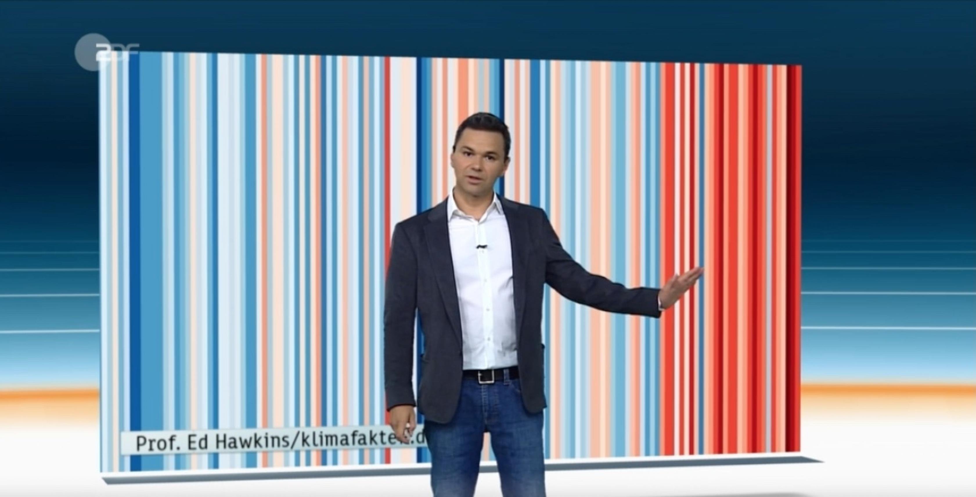 Der ZDF-Wettermoderator Özden Terli steht mit dem Rücken zu einer Streifengrafik und deutet auf ihr vom Zuschauer aus gesehen rechtes Ende. Es handelt sich um die „Warming Stripes“ für Deutschland, bei der für jedes Jahr ein blauer oder roter Streifen zeigt, wie weit die Temperatur vom Mittelwert abwich. Rot überwiegt an der rechten Seite, wo die jüngere Vergangenheit zu sehen ist. Das Bild ist ein Screenshot vom Ende des Heute-Journals am 17. Juli 2018. „Die Effekte des Klimawandels überholen uns“, sagt Terli, „aber wir machen einfach so weiter wie bisher.“