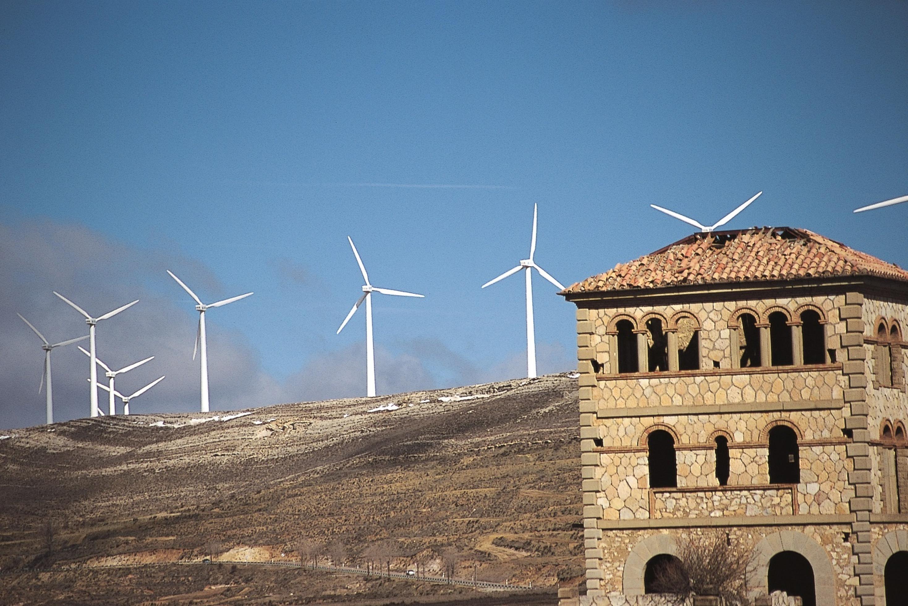 Im Vordergund ein verwaistes Steinhaus, im Hintergrund ein Bergrücken mit Windkraftanlagen.