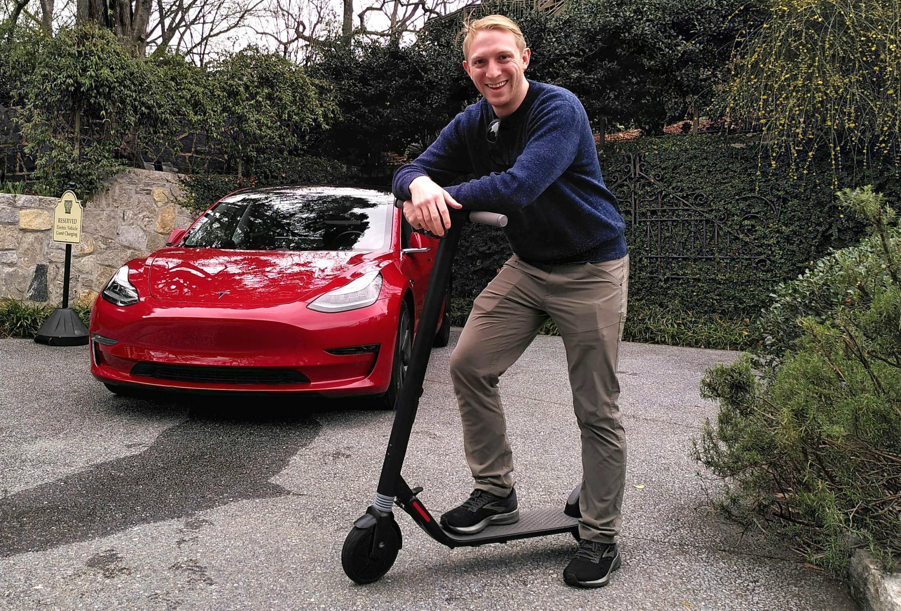 Ein junger Mann steht auf einem E-Scooter. Im Hintergrund steht ein Tesla Model 3.