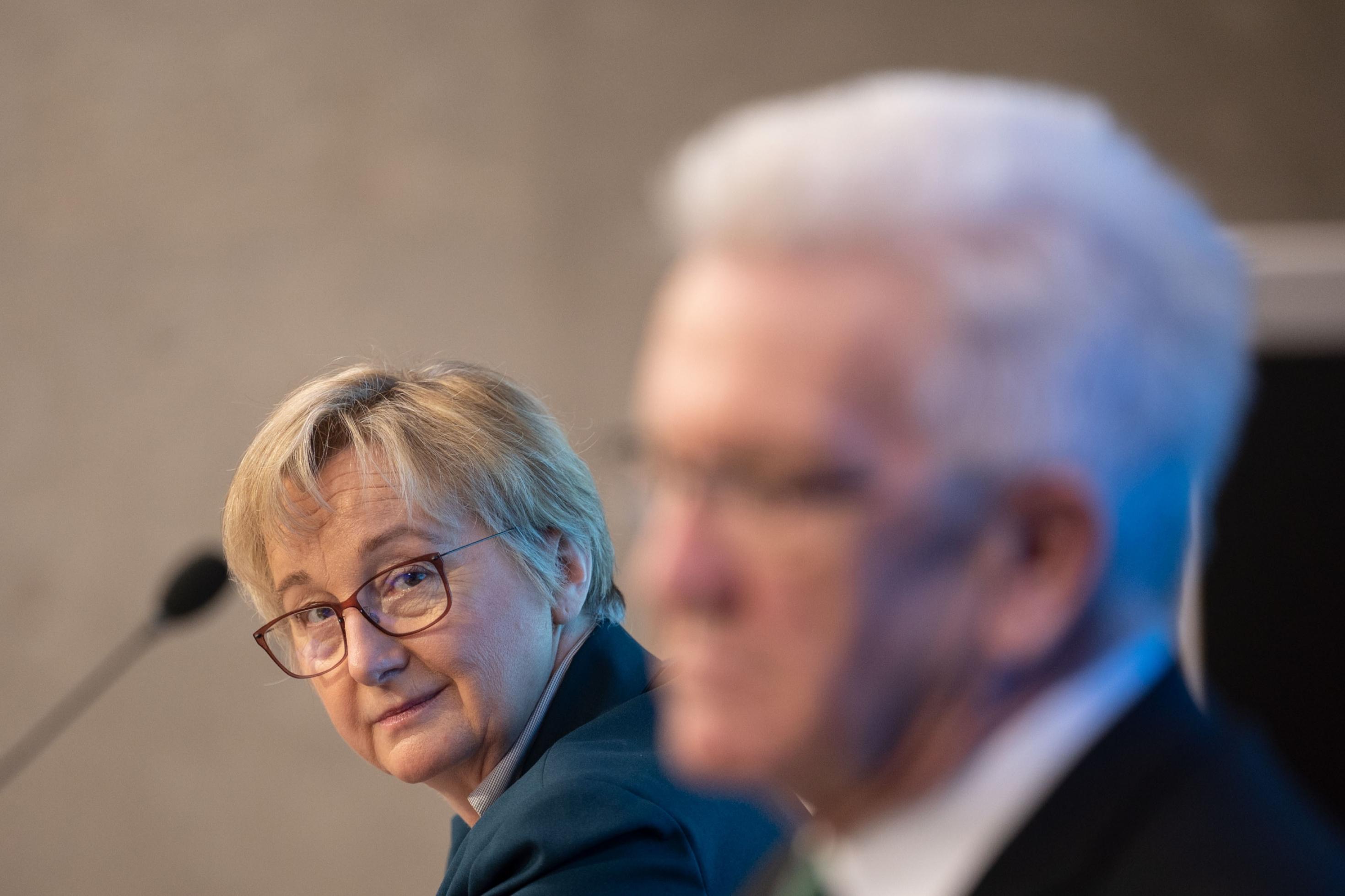 Wissenschaftsministerin Theresia Bauer, eine mittelalte Frau, blond, mit Brille lächelt verschmitzt Winfried Kretschmann zu. Kretschmann ist Ministerpräsident von Baden-Württemberg. Er ist unscharf nur im Profil zu sehen.