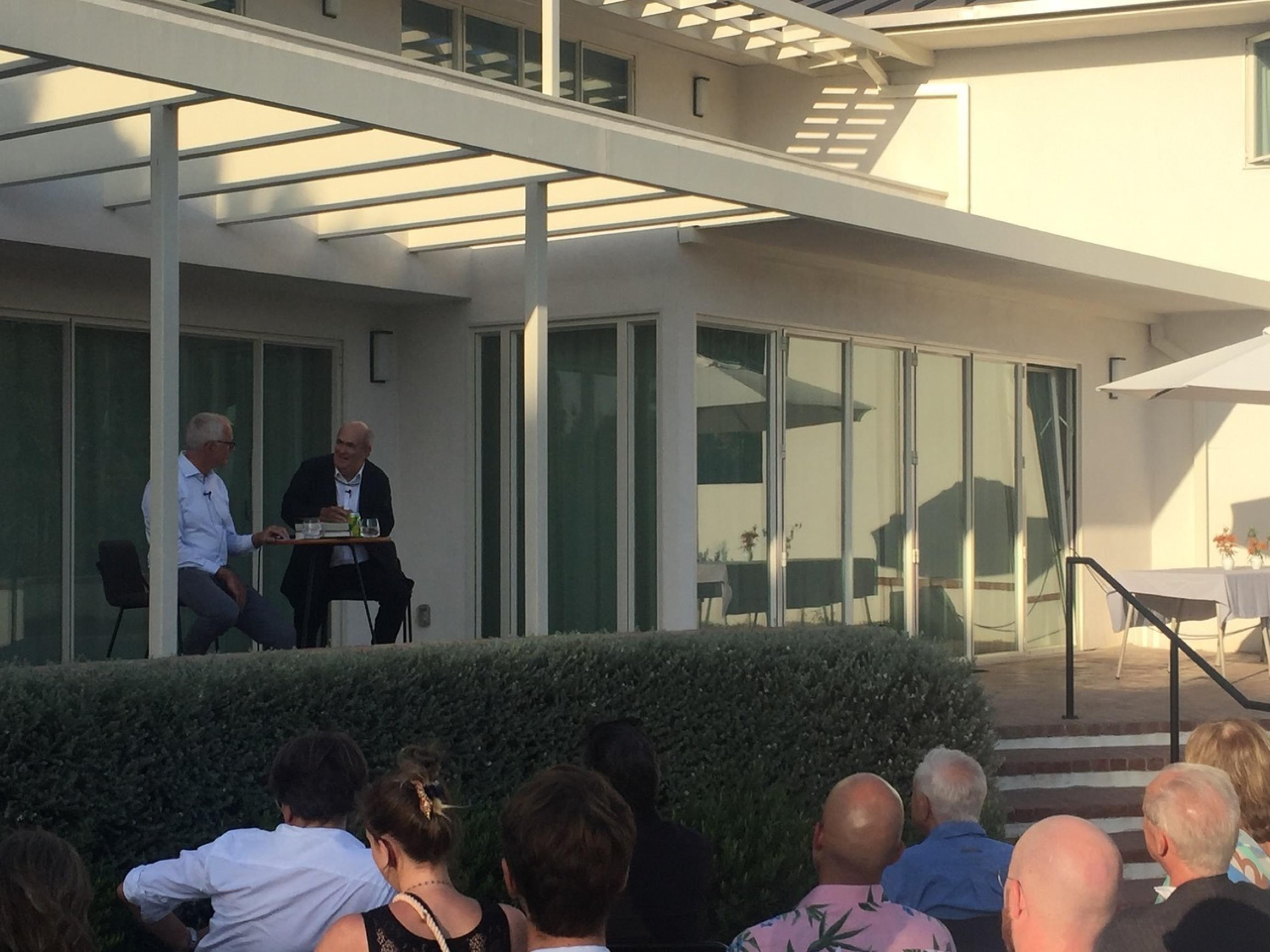 Zwei Männer auf Barhockern an einem runden Bartisch auf einer Terrasse im Sonnenlicht. Vor ihnen eine Hecke und davor Zuschauer, die ihnen aufmerksam zuhören.