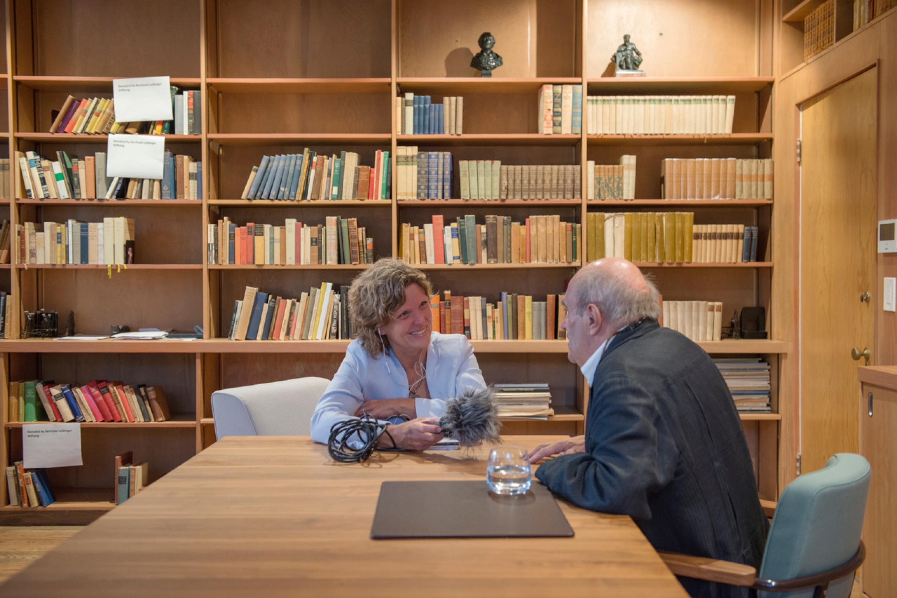 Eine Frau in hellblauem Blazer interviewt einen Herrn mit grauem Haar, Rücken zum Fotografen, an einem großen Schreibtisch. im Hintergrund Bücherregale.