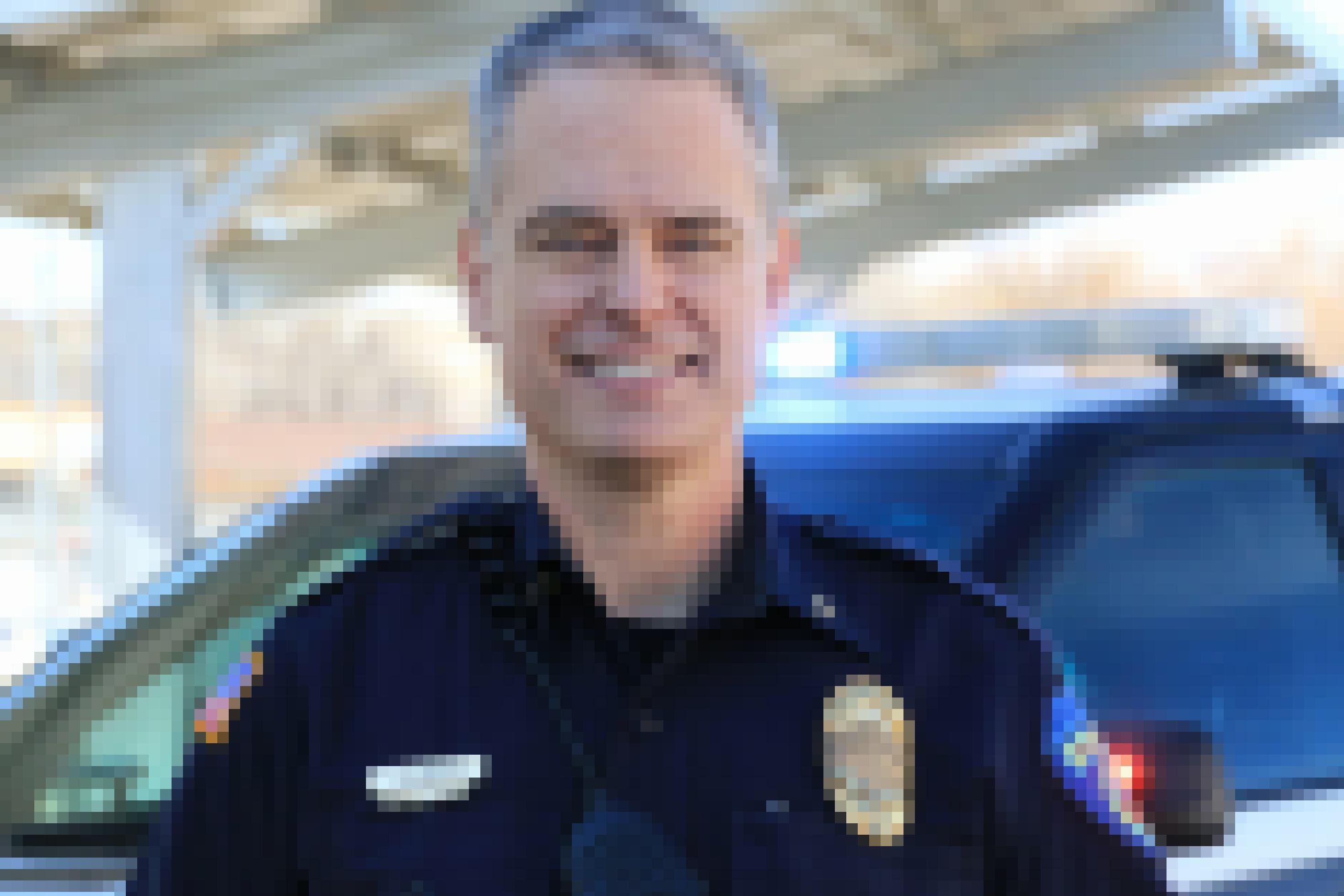 Ein Polizist in Uniform lächelt in die Kamera. Im Hintergrund steht ein Einsatzwagen mit angeschalteten Signalleuchten.