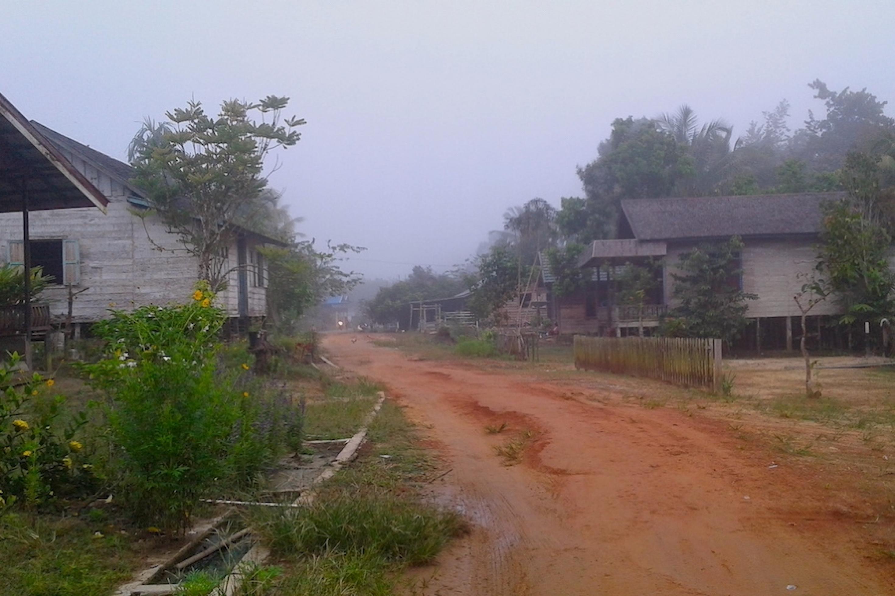 Ungepflasterte Dorfstraße von Tumbang Mantuhe im dunstigen Licht der Dämmerung führt entlang einfacher Holzhäuser auf Stelzen