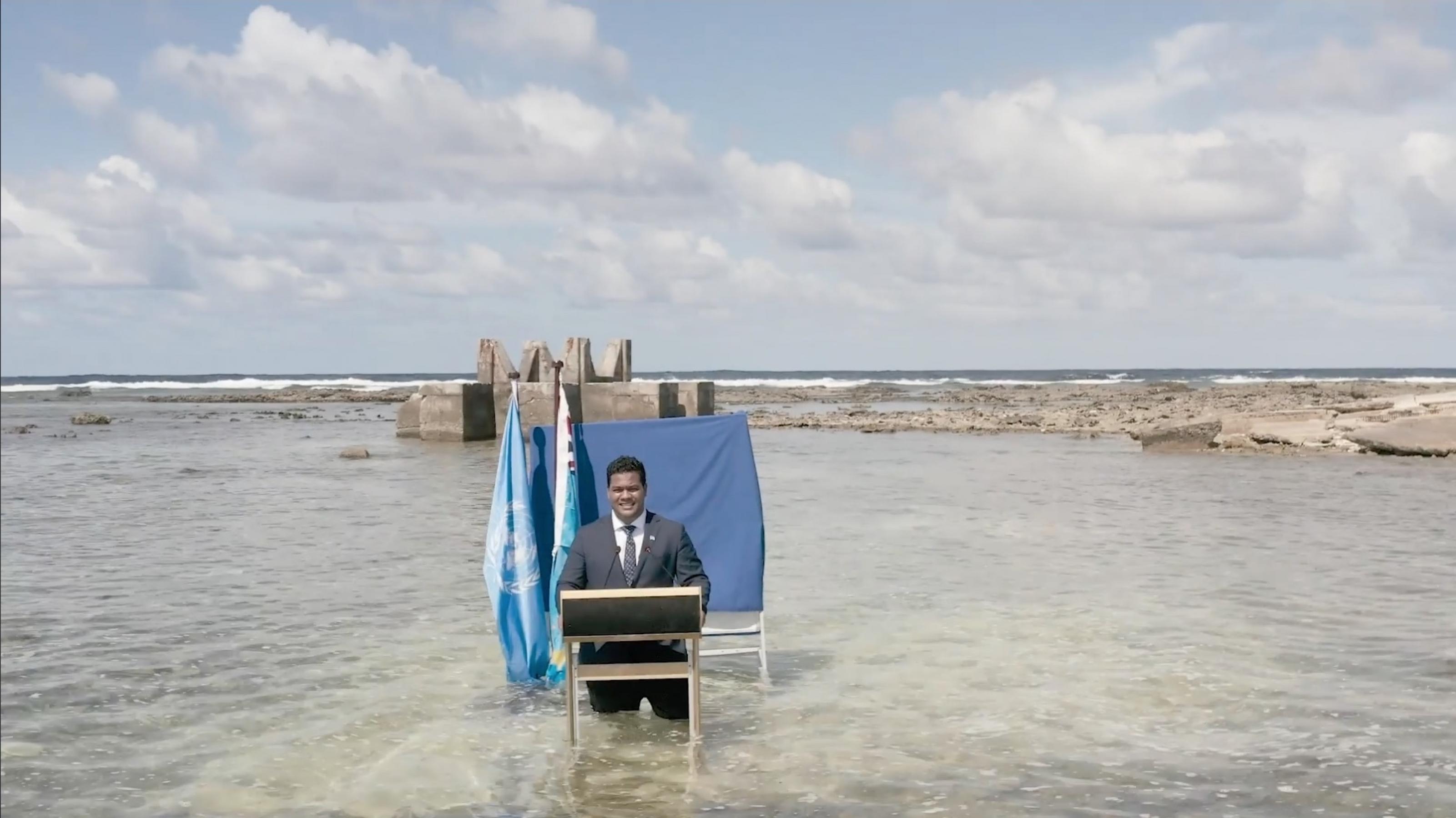 Der Politiker steht an einem Rednerpult vor einem dunkelblauen Vorhang und zwei Flaggen. Das Wasser einer Lagune umspült seine Beine. Im Hintergrund ist eine Gebäuderuine zu sehen. Das Bild ist von einer Drohne aus einigen Metern Höhe aufgenommen.