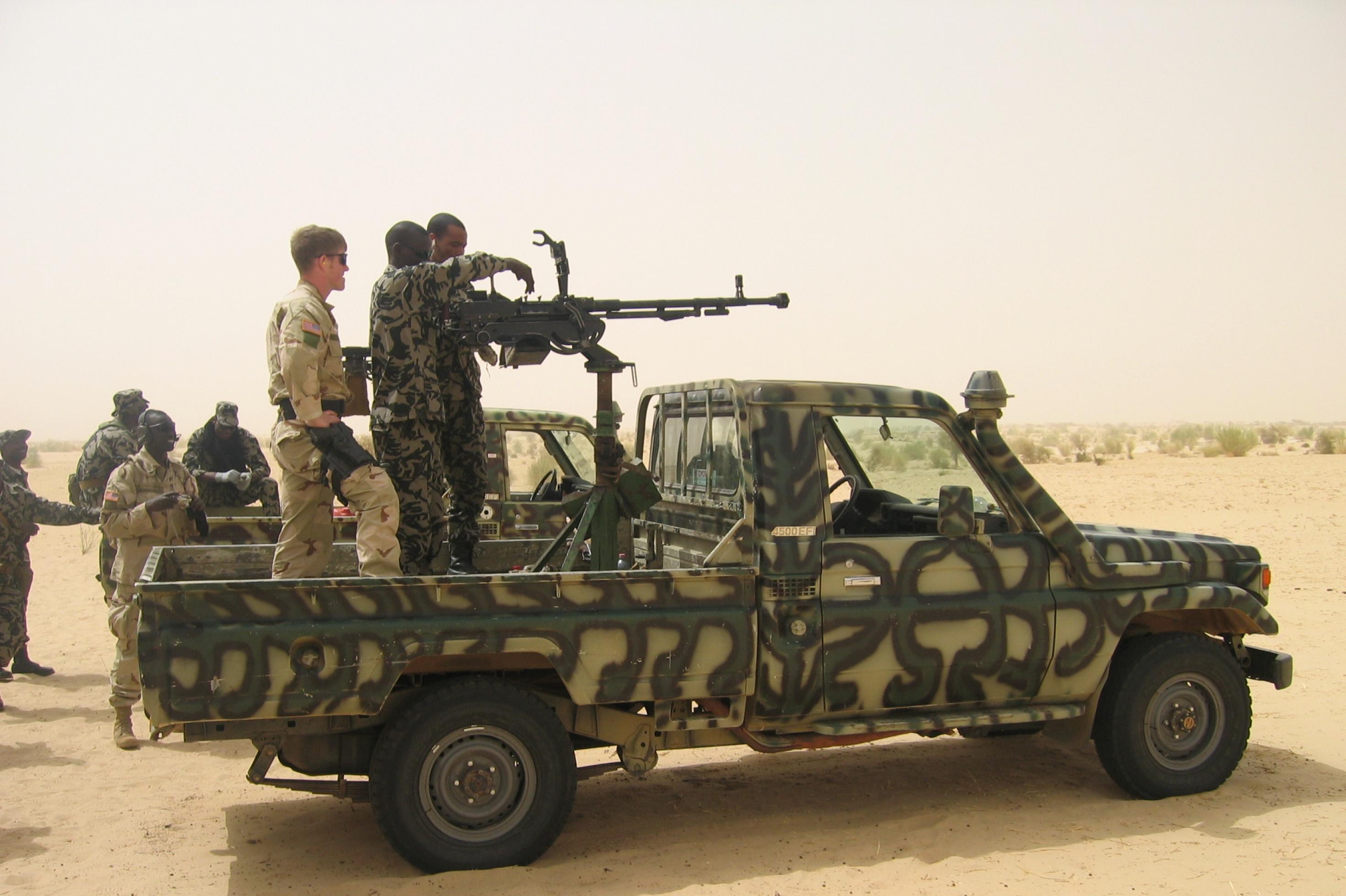 Zu sehen ist ein Pick-Up in militärgrün, in Tarnfarben. Auf der Ladefläche ist ein Geschütz aufmontiert, die Luft ist von Sand verhangen. Am Geschütz stehen US-amerikanische und malische Soldaten – offensichtlich eine Trainingssituation.