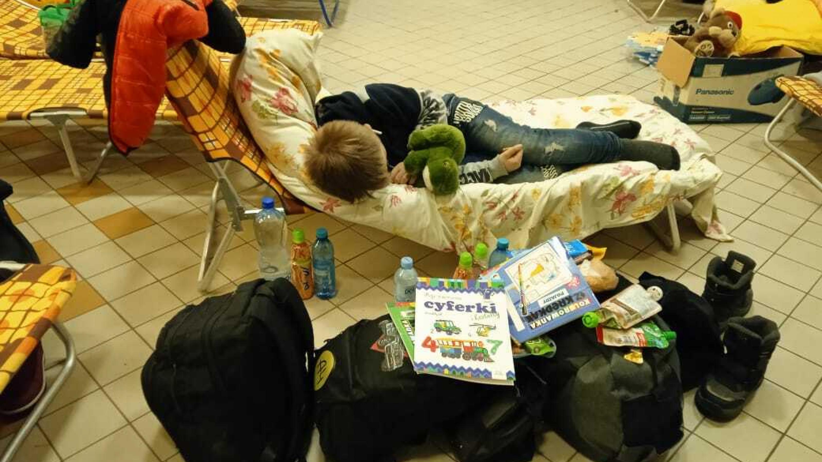 Ein Junge schläft auf einer Liege, vor ihm das Gepäck der Flüchlingsfamilie. Auf den Taschen liegen Kinderbücher.