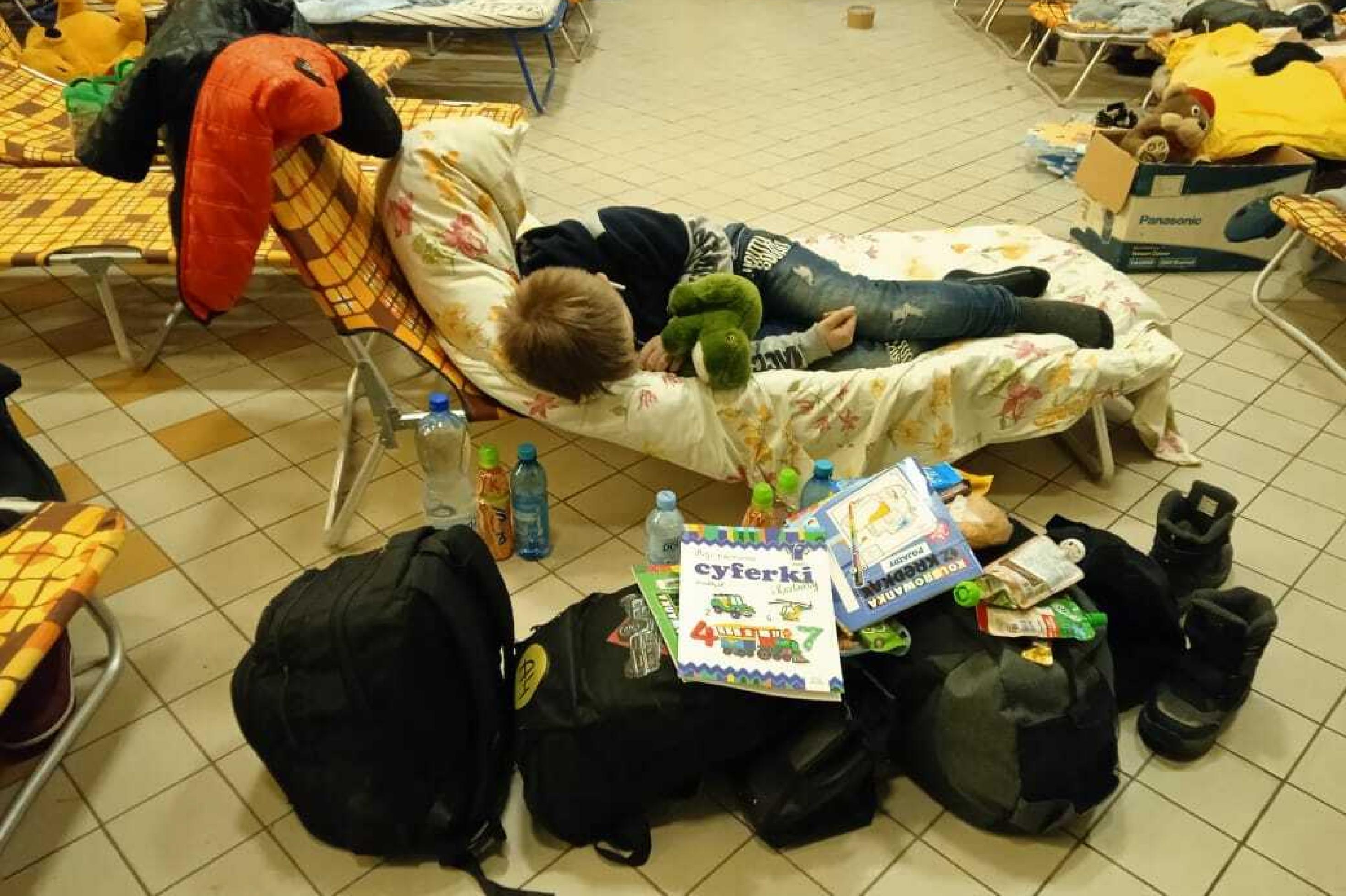 Ein Junge schläft auf einer Liege, vor ihm das Gepäck der Flüchlingsfamilie. Auf den Taschen liegen Kinderbücher.