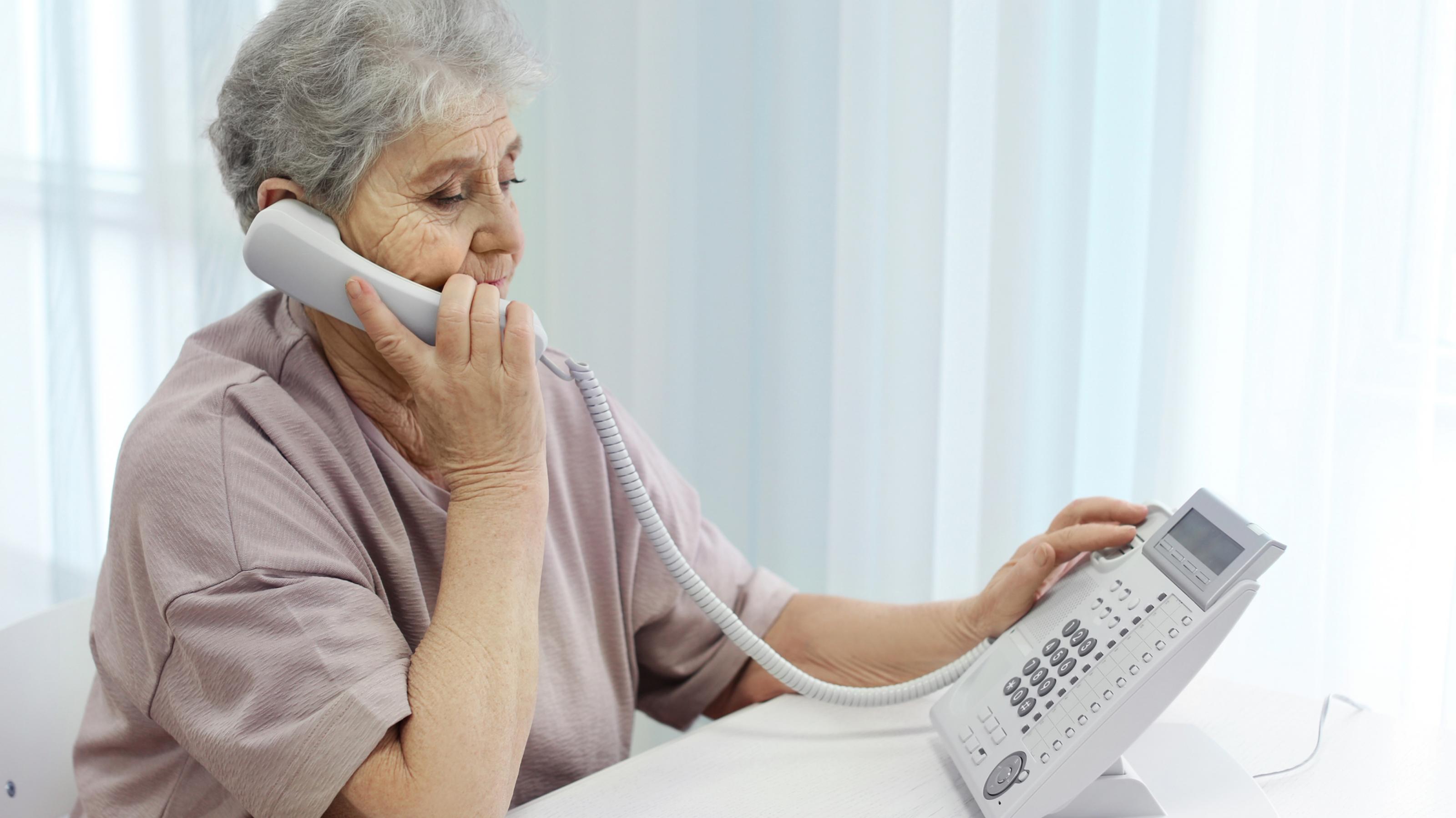 Ältere Dame sitzt vor einem Telefon, hält einen Hörer ans Ohr wählt