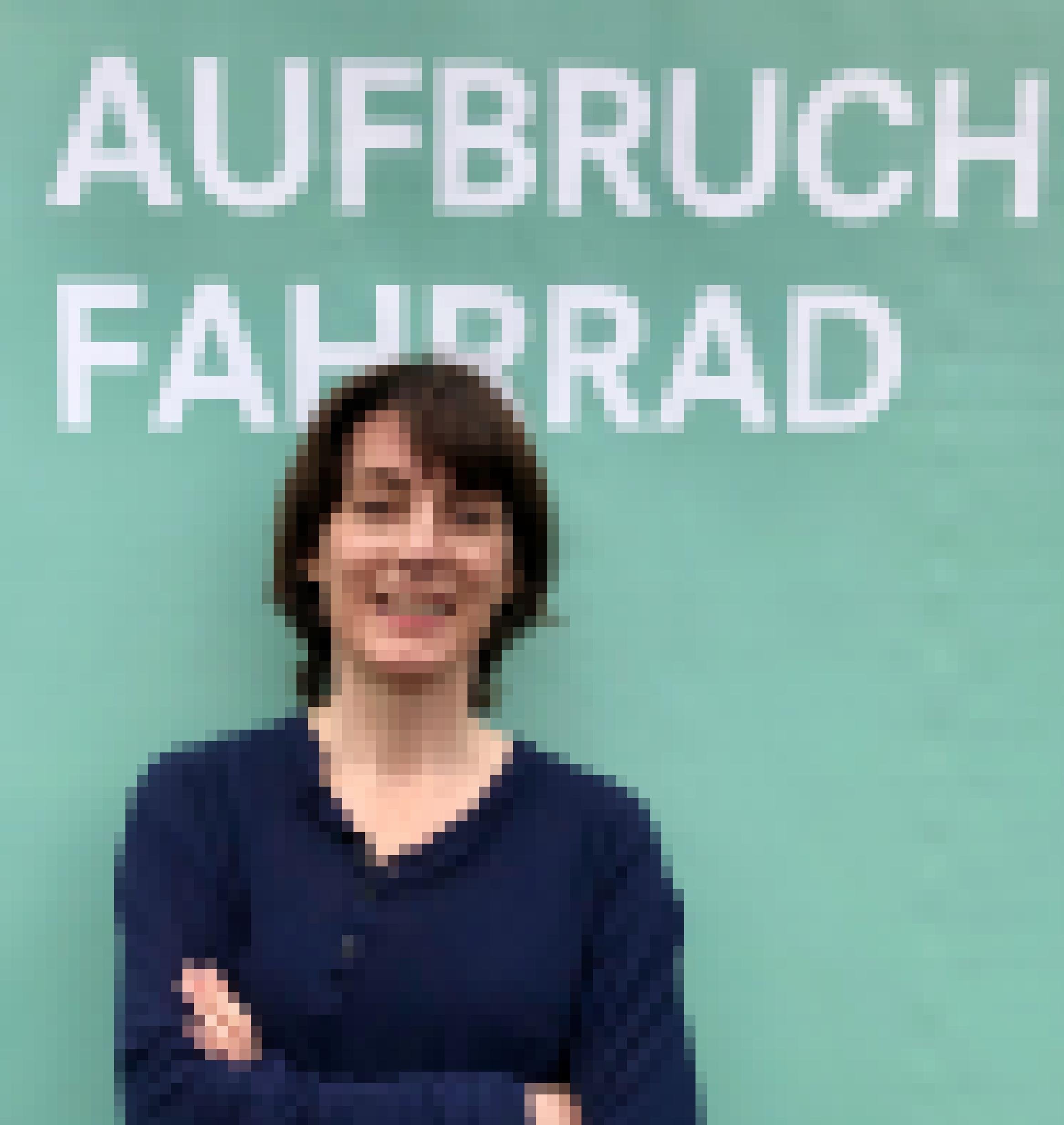 Ute Symanski hat kurzes, schulterlanges Haar, sie trägt ein dunkelblaues langärmeliges T-Shirt mit drei Knöpfen und lacht in die Kamera. Sie lehnt an einer türkisfarbenen Wand mit dem weißen Schriftzug „Aufbruch Fahrrad“