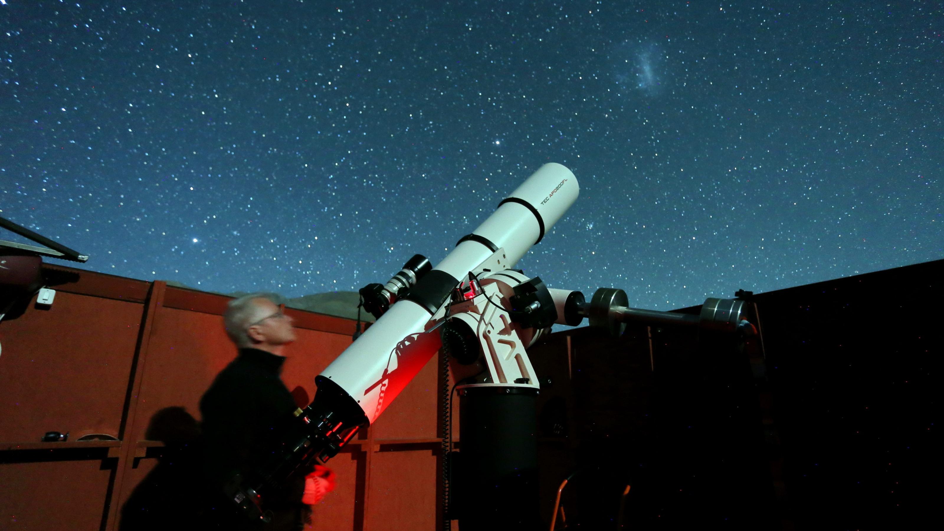 Ein Mann steht neben einem großen Fernrohr in einer Schutzhütte mit geöffnetem Dach und fotografiert den nächtlichen Sternenhimmel