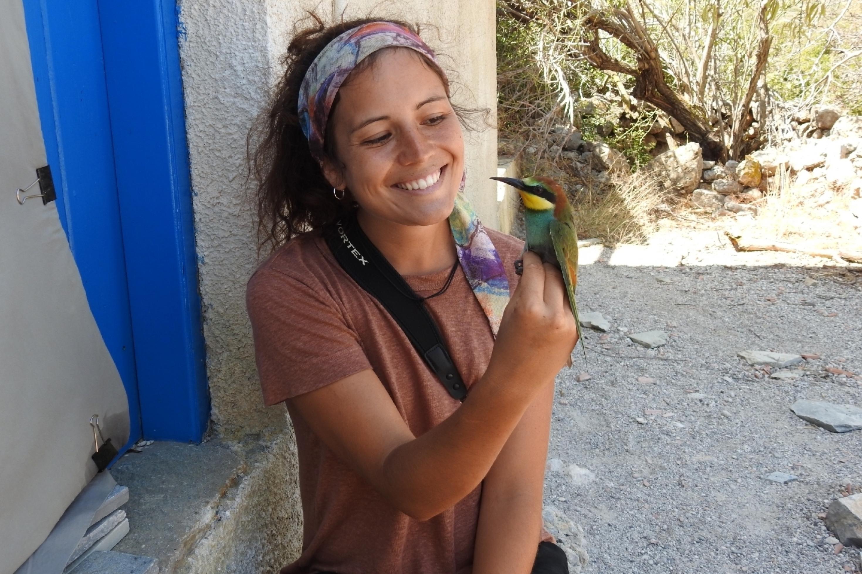 Die junge Ornithologin Verena Rupprecht sitzt vor einer Hütte und hält einen bunten Vogel in der Hand, einen Bienenfresser