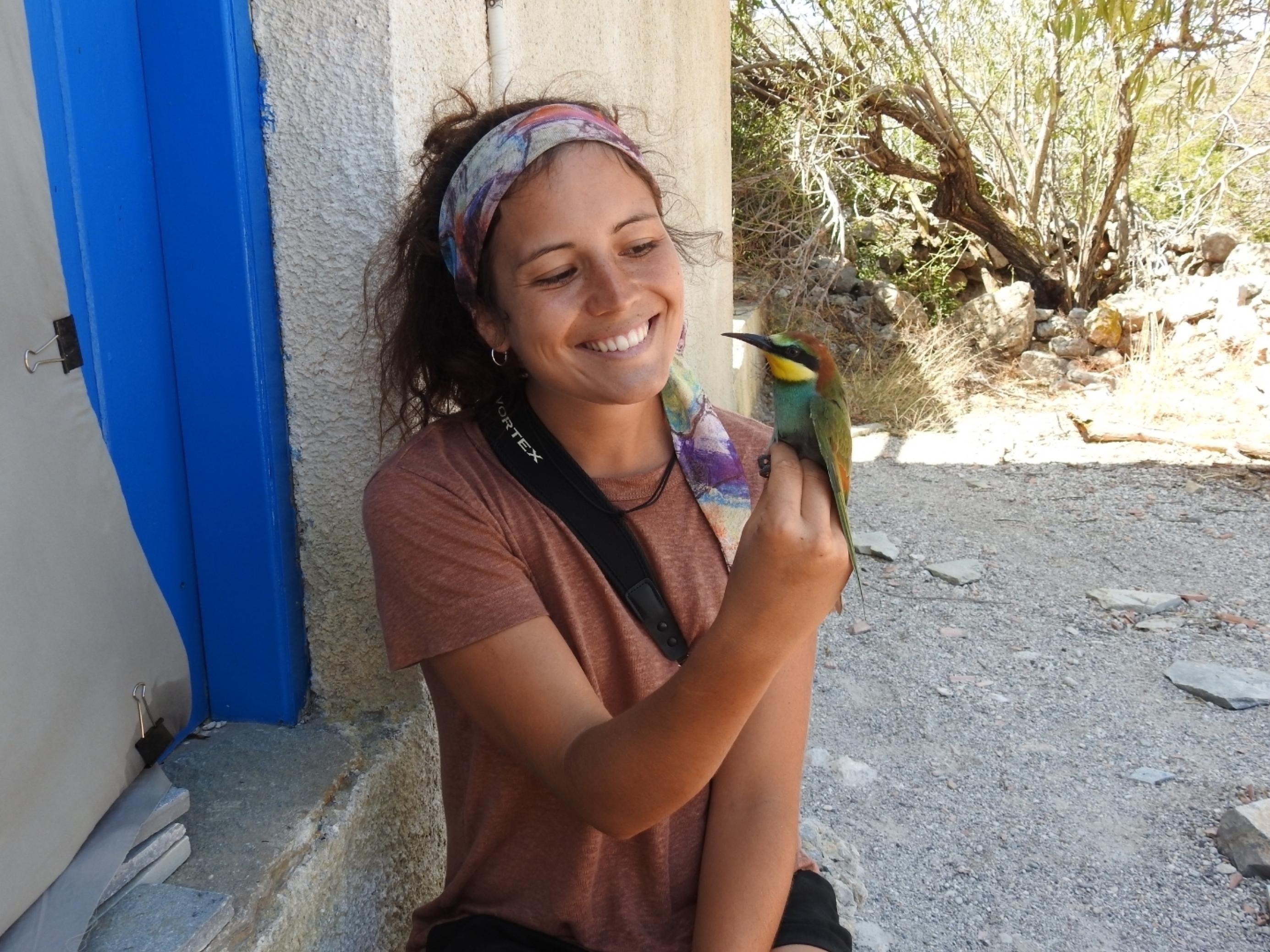 Die junge Ornithologin Verena Rupprecht sitzt vor einer Hütte und hält einen bunten Vogel in der Hand, einen Bienenfresser