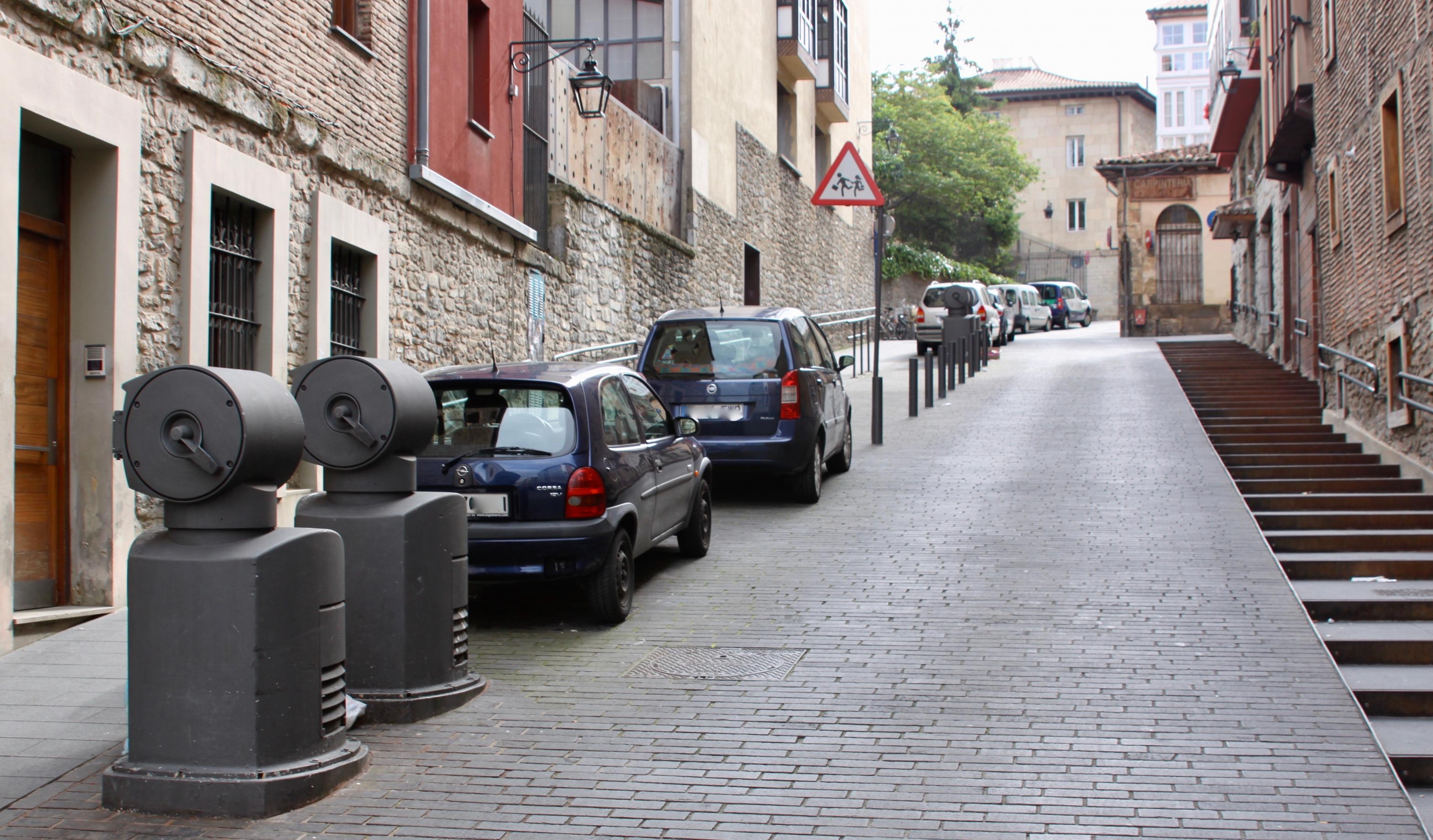 Eine kleine Gasse. Links stehen vor zwei Müllschluckern parkende Autos.