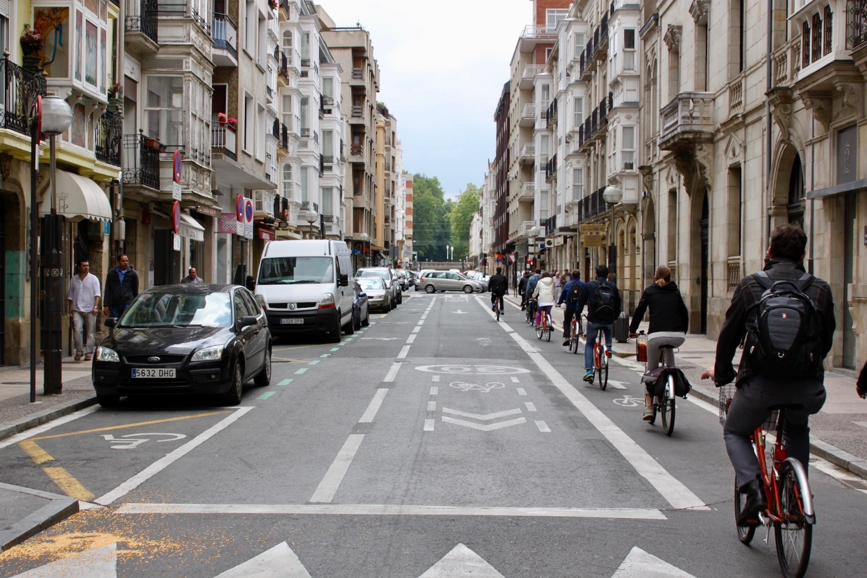Eine Straße, die zwischen Altbauten verläuft. Radfahrer nutzen ein Teil der rechten Spur, links stehen parkende Autos.