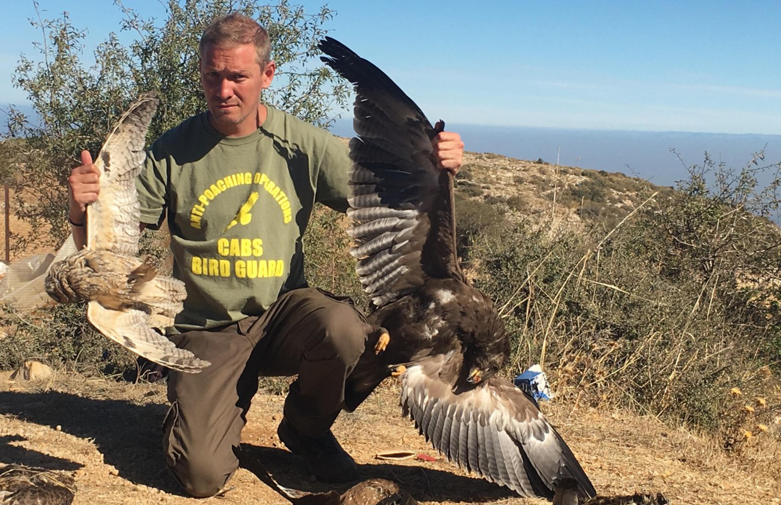 Lloyd Scott kniet auf dem Boden und hält dabei zwei getötete Vögel in die Luft: einen Schreiadler und einer Waldohreule.