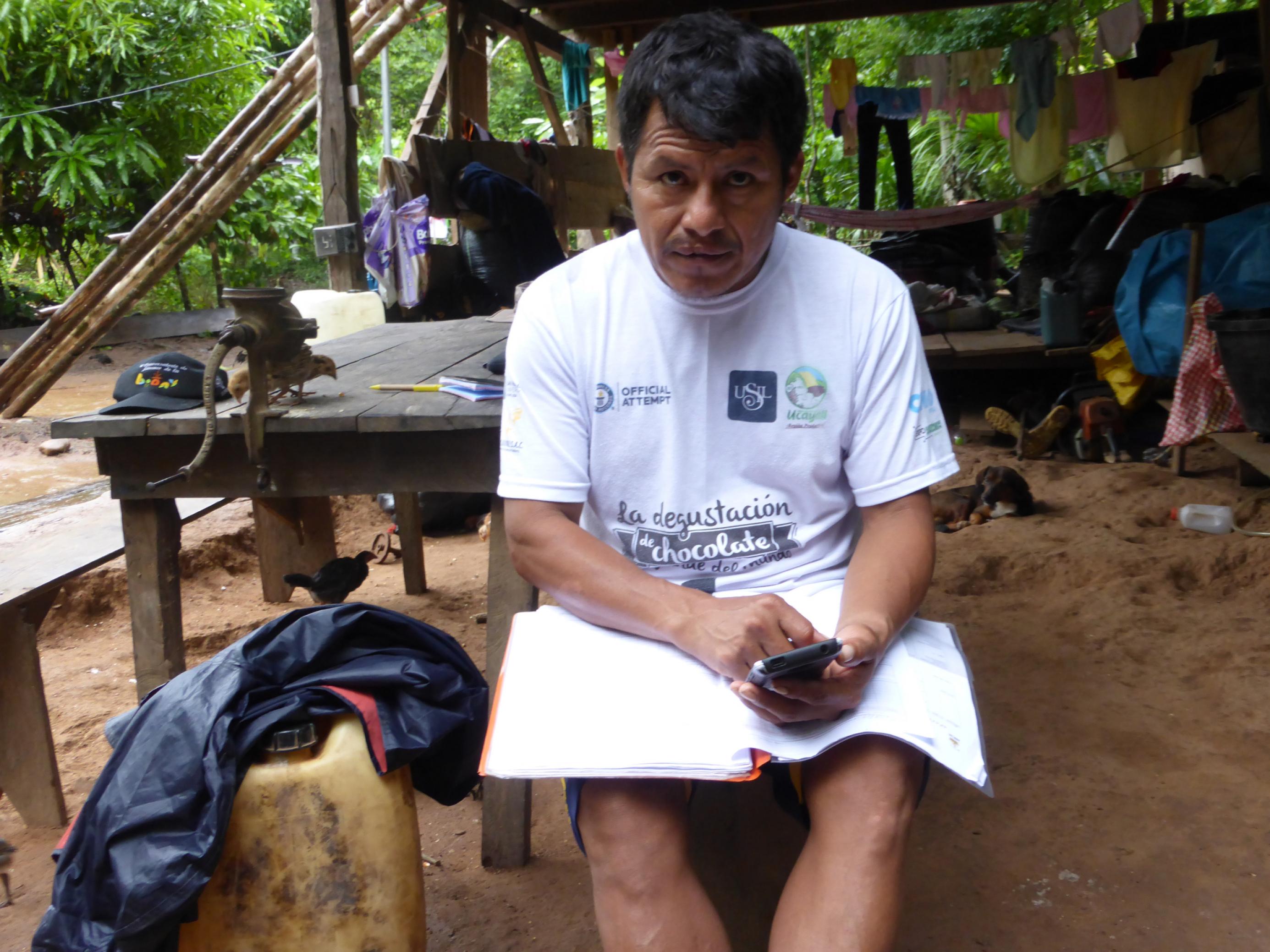 Ein indigener Mann in weissem T-Shirt sitzt auf einer Holzbank und tipp etwas in ein Handy.
