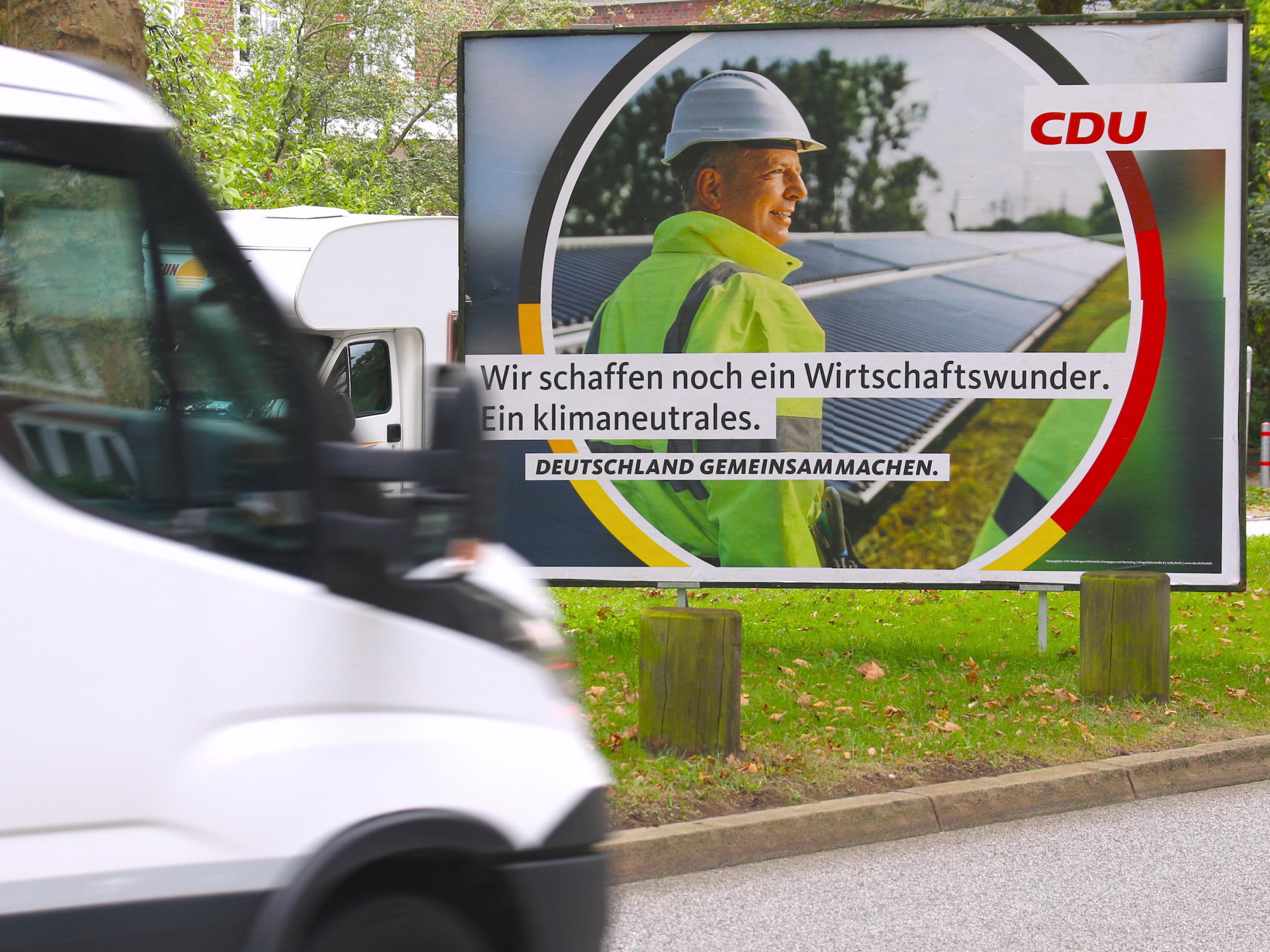 Ein Wahlplakat der CDU zeigt einen Mann mit Bauarbeiterhelm und neongelbem Anorak vor einer Reihe von Solarpanels. Der Slogan lautet: „Wir schaffen noch ein Wirtschaftswunder. Ein klimaneutrales.“ Das Plakat steht auf einem Grünstreifen an der Straße. Von links fährt ein weißer Kleinlaster ins Bild.