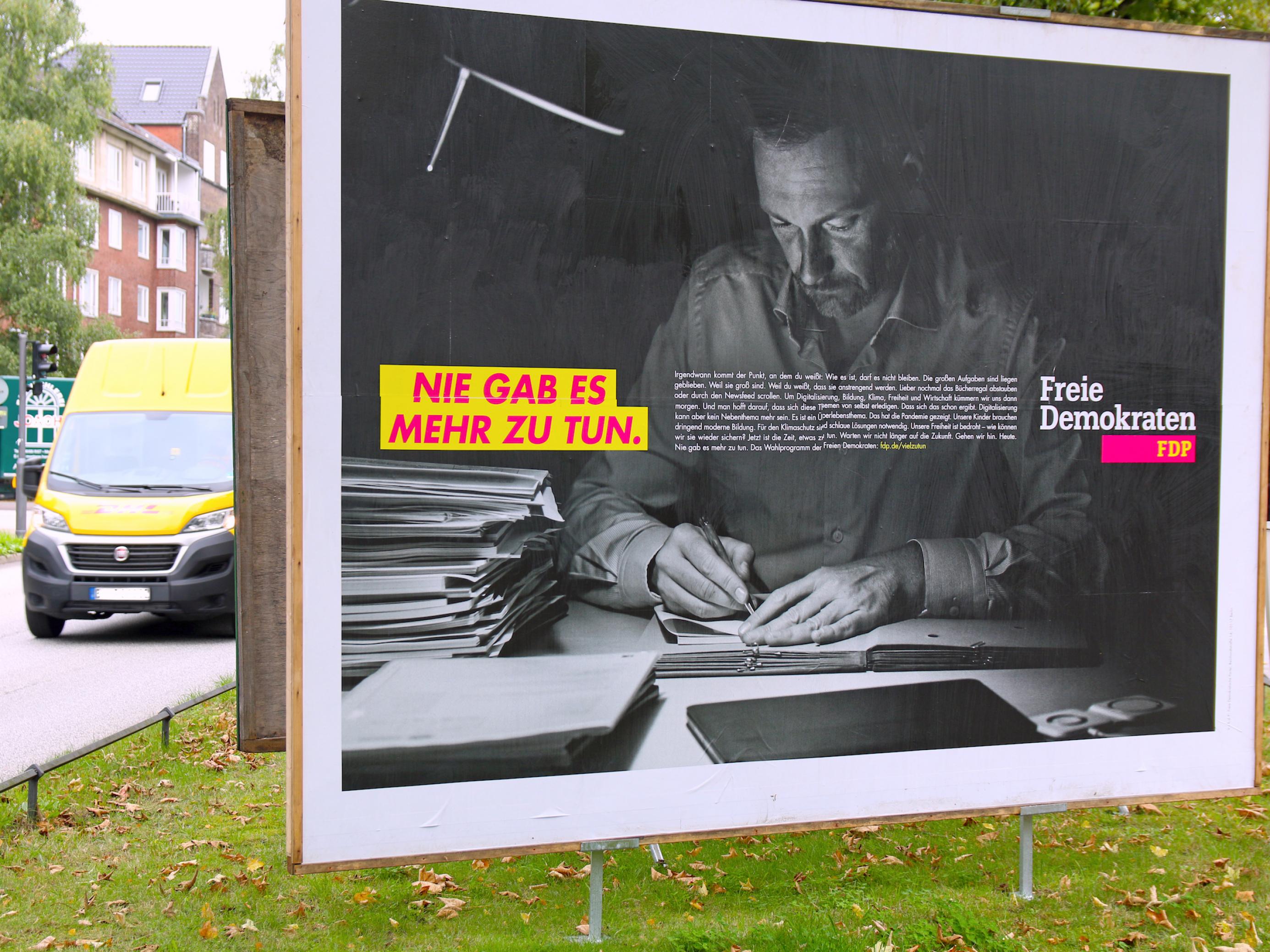 Wahlplakat der FDP: Partei-Chef Christian Lindner sitzt nachts am Schreibtisch und macht Notizen in einer Kladde. Neben ihm auf dem Schwarz-Weiß-Foto liegt ein Stapel Aktendeckel. Der Slogan in geblockten Buchstaben auf gelbem Grund ist: „Nie gab es mehr zu tun“. Das Plakat steht auf einem Grünstreifen, links fährt ein gelber Lieferwagen vorbei.
