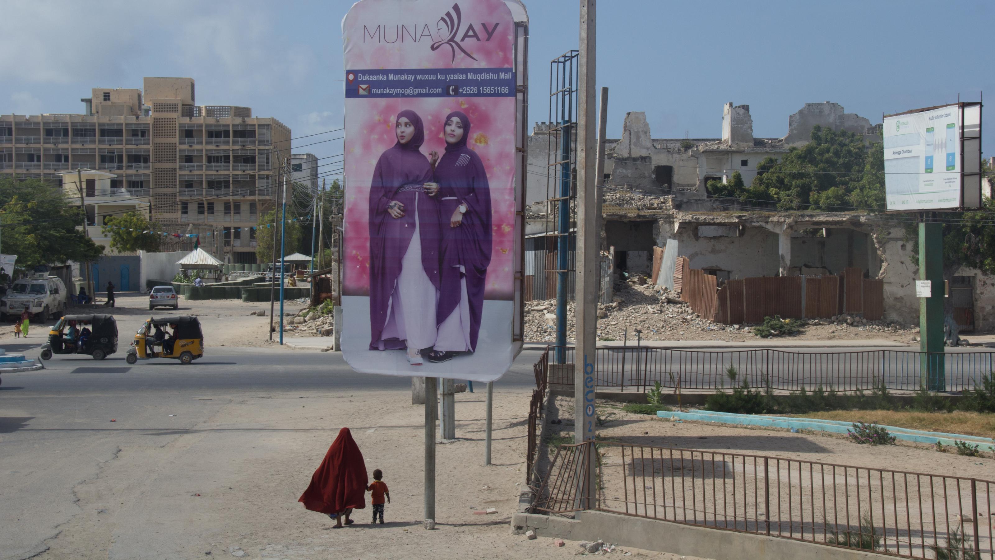 Zu sehen ist ein riesiges Werbeplakat mit zwei Frauen, die Kopftuch tragen. Darunter winzig klein von hinten eine Frau mit Kopftuch, an der Hand ein Kind. Im Hintergrund Ruinen.