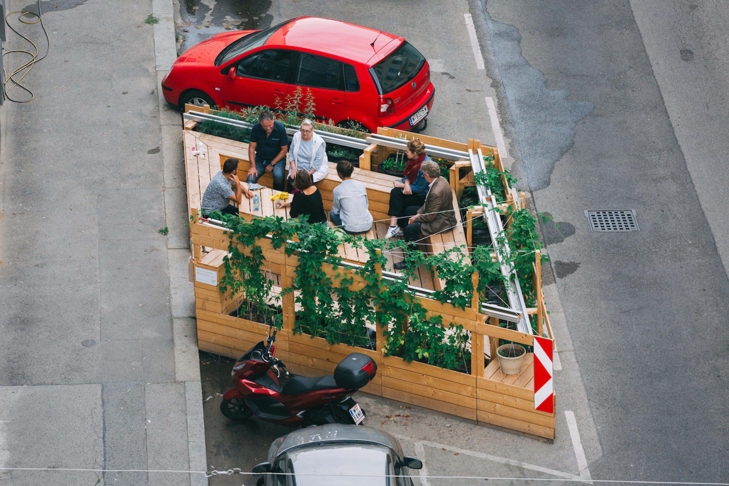 Blick aus der Vogelperspektive auf eine Holzterrasse, die auf einen Parkplatz aufgebaut wurde. Dort sitzen sechs Menschen auf den Holzbänken und unterhalten sich