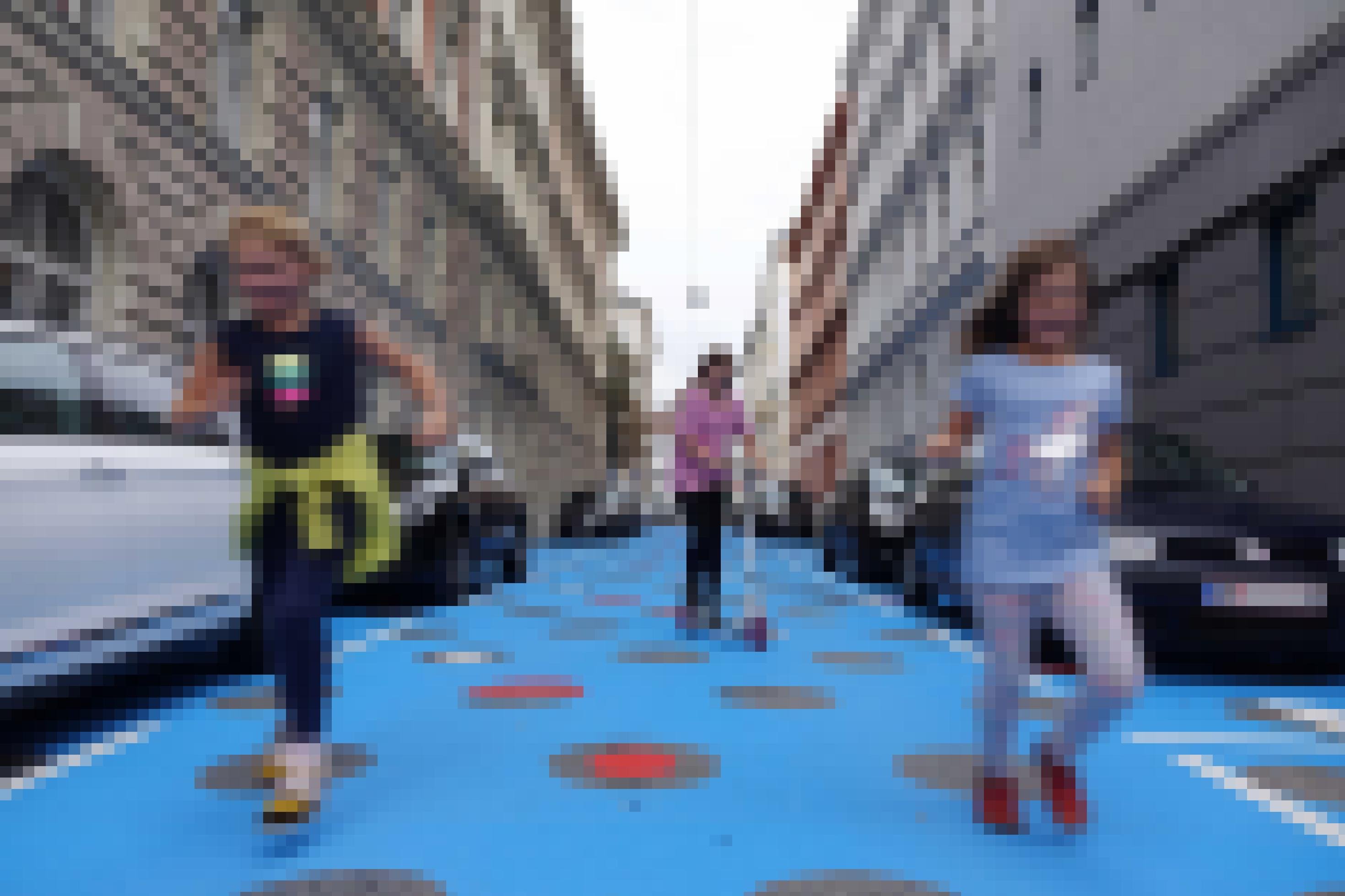 Der Asphalt in der Wohnstraße ist blau gestrichen worden und hat rote, weiße und asphaltfarbene Kreise. Kinder laufen durch die Straße oder fahren Roller. Rechts und links am Fahrbahnrand parken Autos