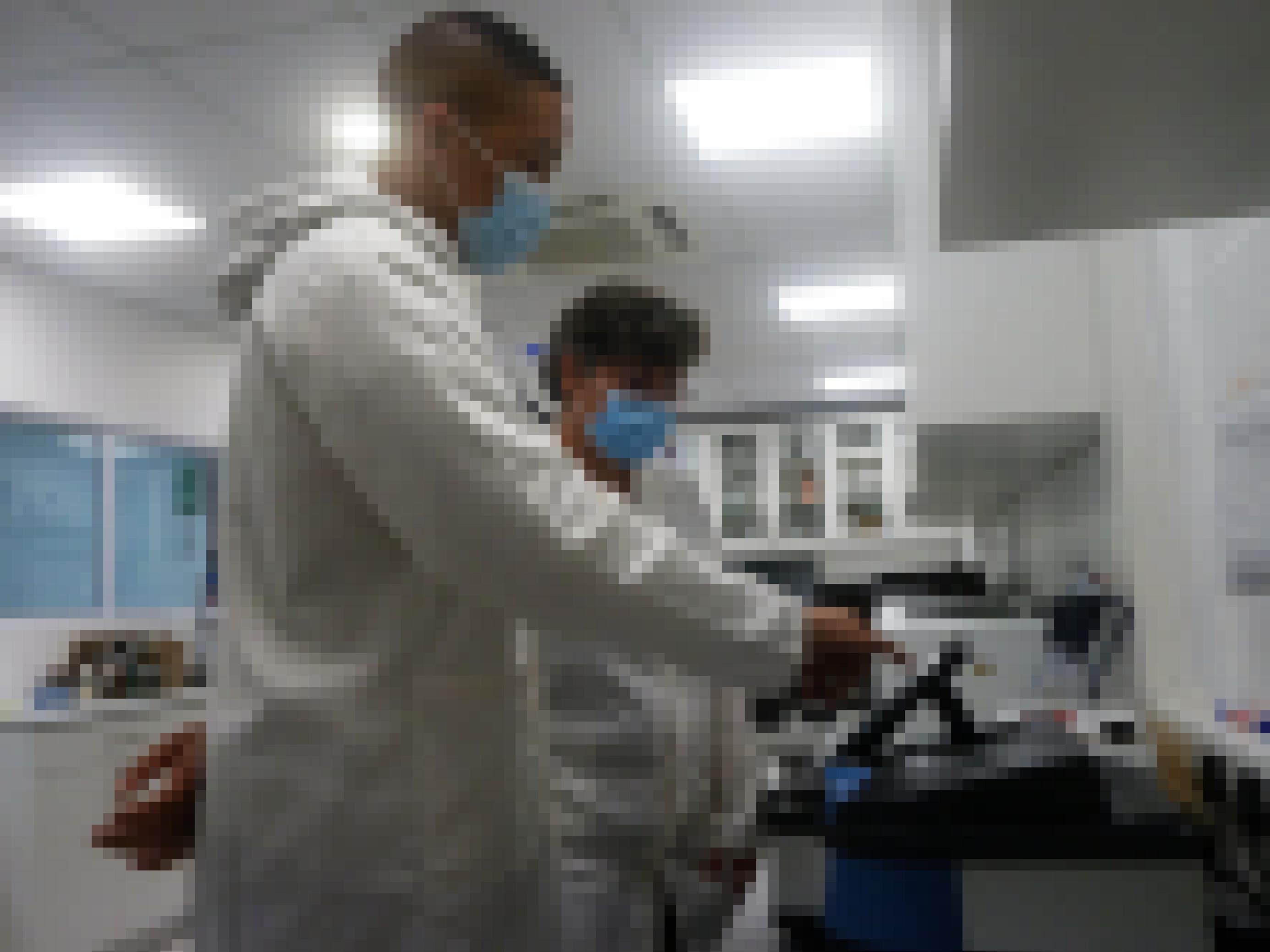 Die beiden stehen in weißen Kitteln und mit OP-Masken im Labor. Der Kollege der Wissenschaftlerin deutet auf einen Bildschirm.