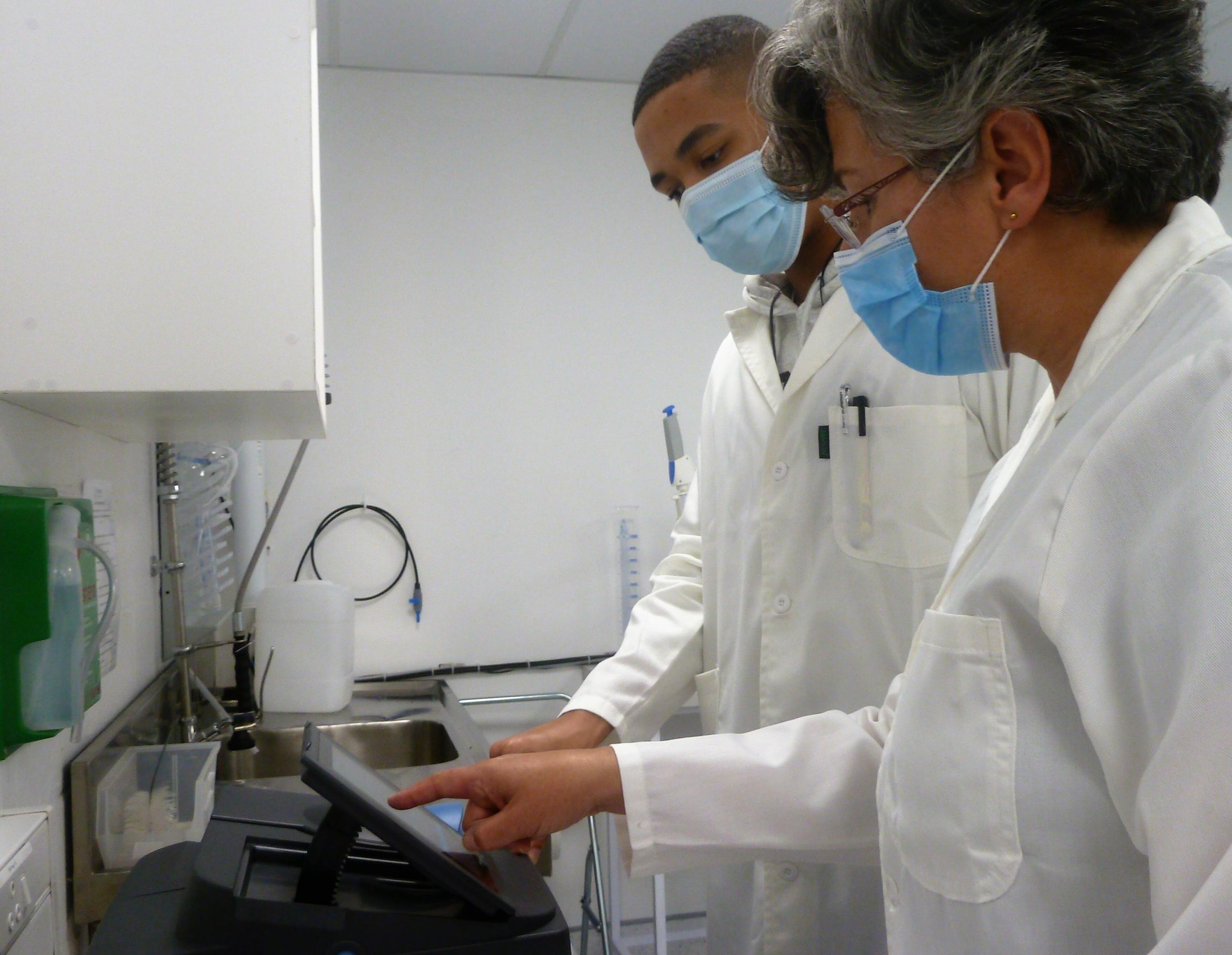 Zwei Forschende mit Masken, eine Frau und ein Mann, schauen im Labor auf die Anzeige eines Labor-Geräts