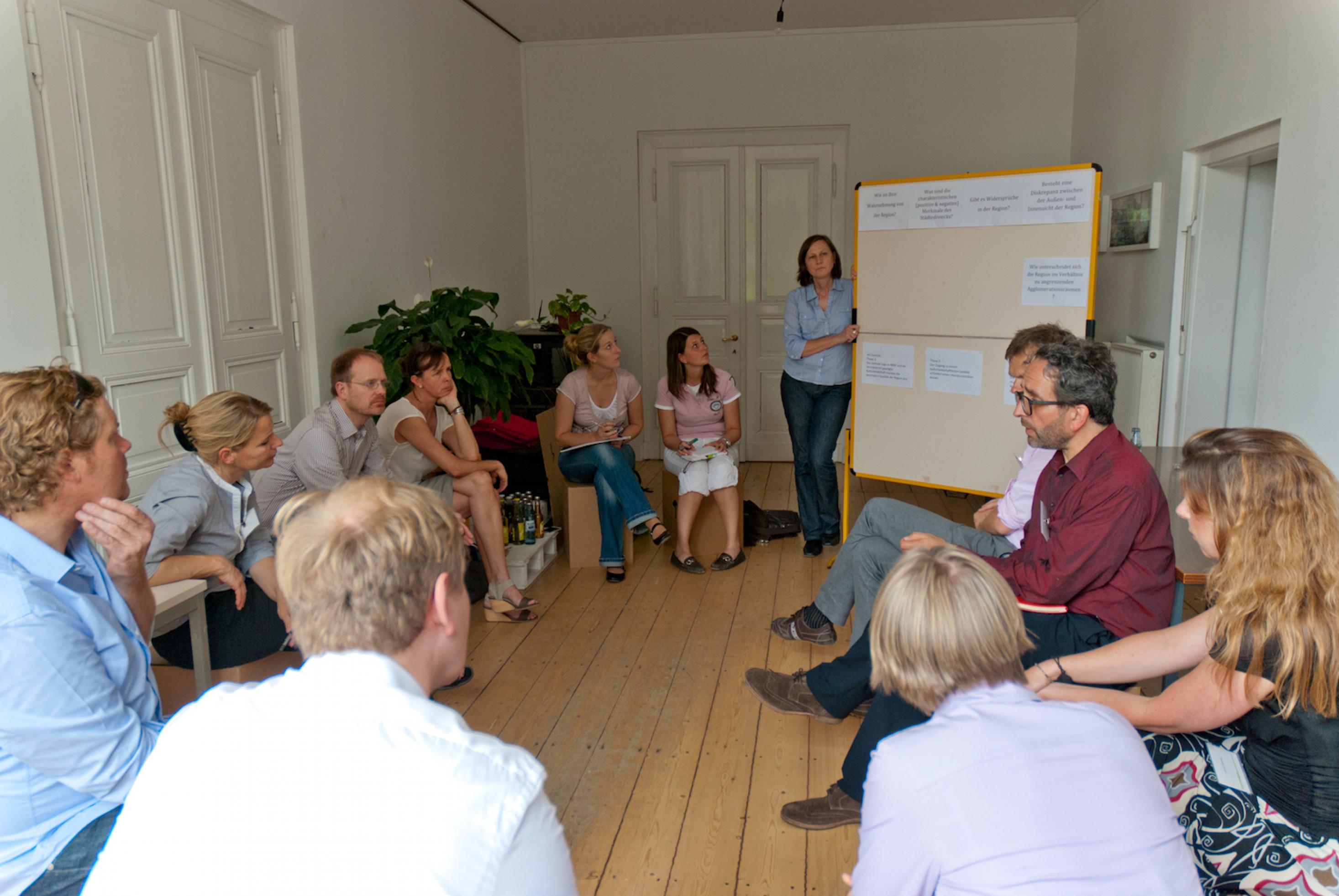 Teilnehmer des Workshops in einem kleinen Raum mit weißen Wänden und hellem Bretterboden.