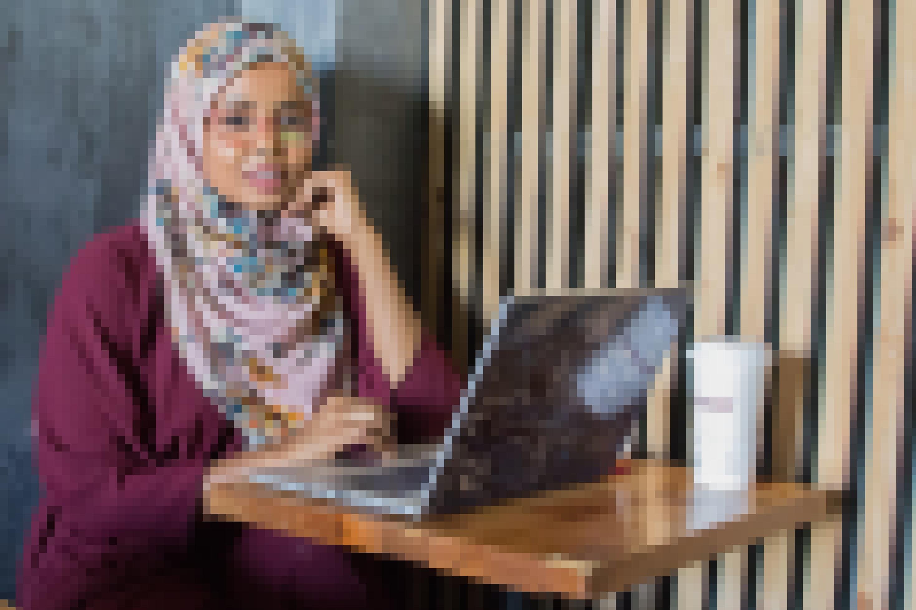 Eine junge Somalierin, freundlicher Blick, mit Laptop und Kaffee im Café, an einem modernen Holztisch.