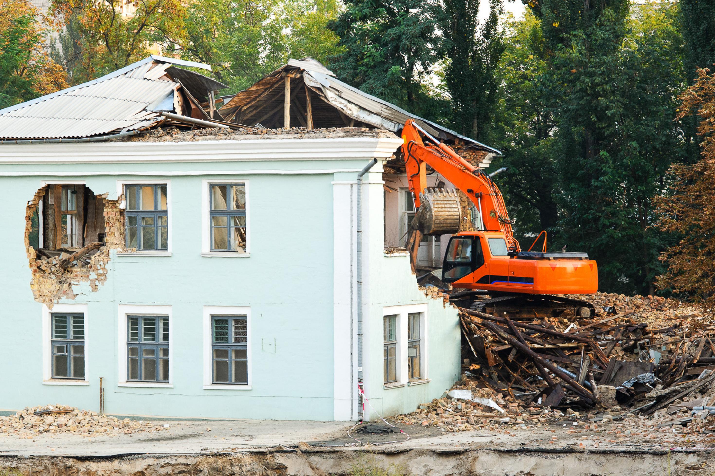 Ein orangefarbener Löffelbagger steht auf einem Haufen Bauschutt neben einem gründerzeitlichen Haus, dem eine ganze Seitenwand, große Teile des Daches und einige Fenster fehlen.