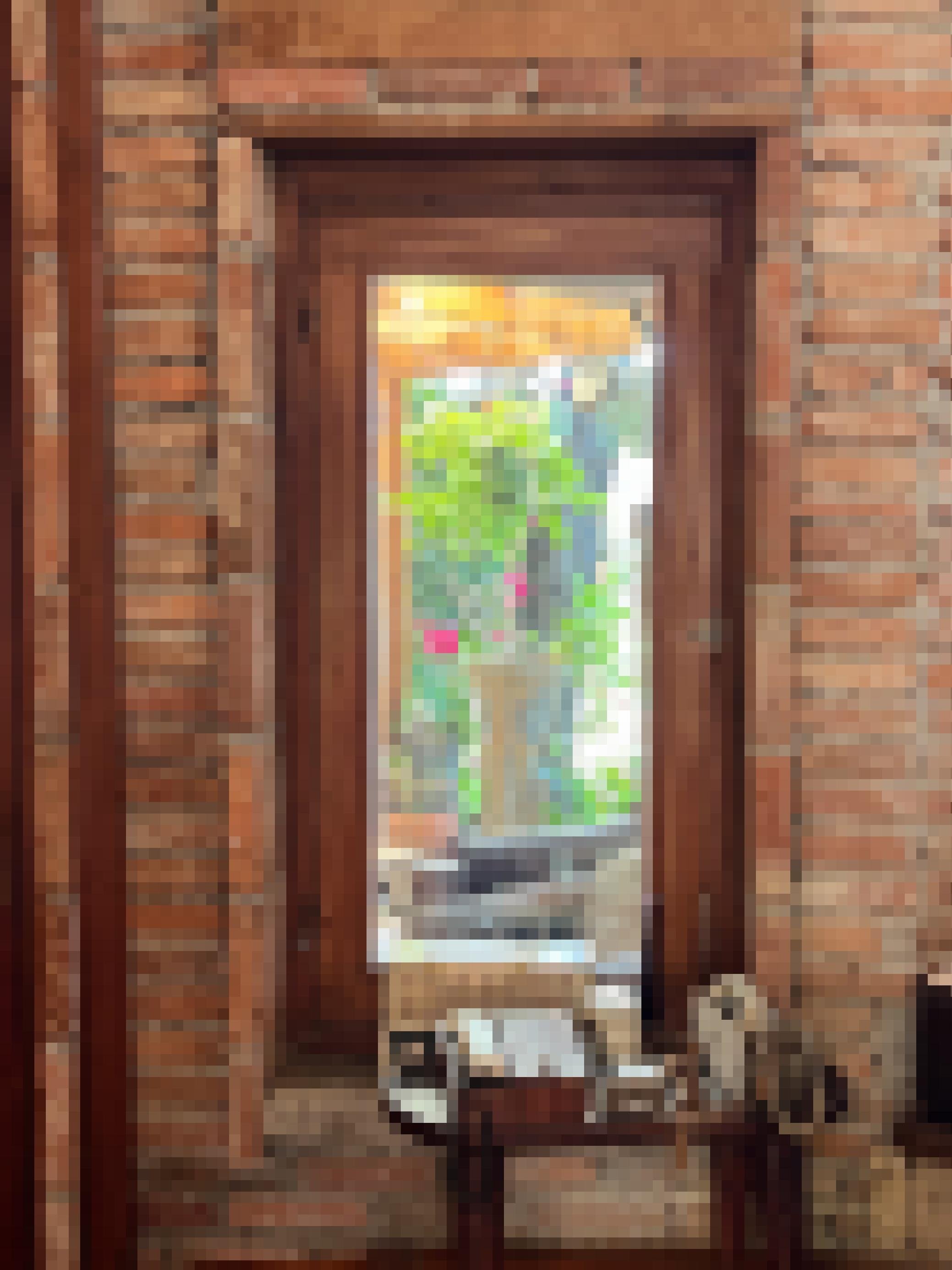 Zu sehen sind unverputzte Ziegelsteine, darin einer hölzerner Fensterrahmen. Der Blick geht auf einen Pflanztopf im Garten mit einer blühender Pflanze, dahinter ein Busch. Im Vordergund ein hölzernes Telefontischchen, auf dem zwei sehr große, vermutlich historische Festnetztelefone stehen.